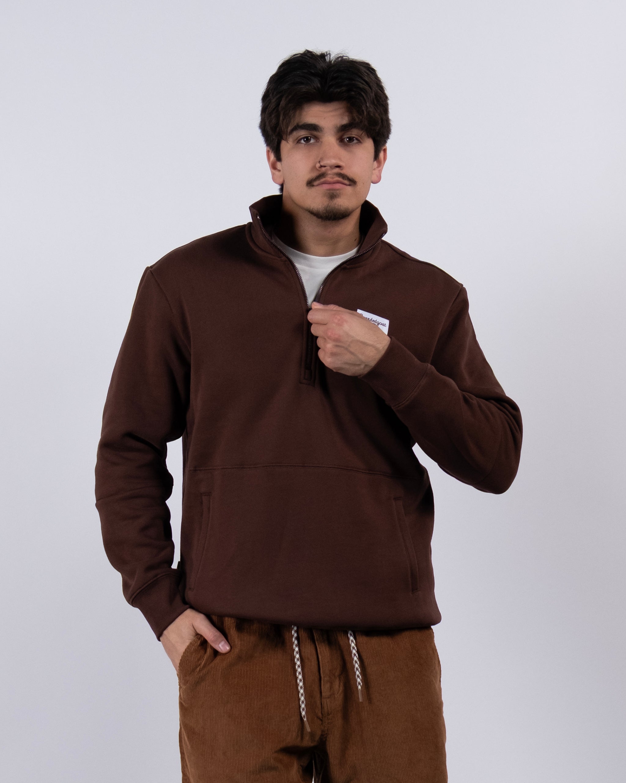 A young man with dark hair and a mustache wears the BEARDEDGOAT NEW — VOID™ Half Zip and brown drawstring pants, standing against a light gray background with one hand in his pocket and a neutral expression.