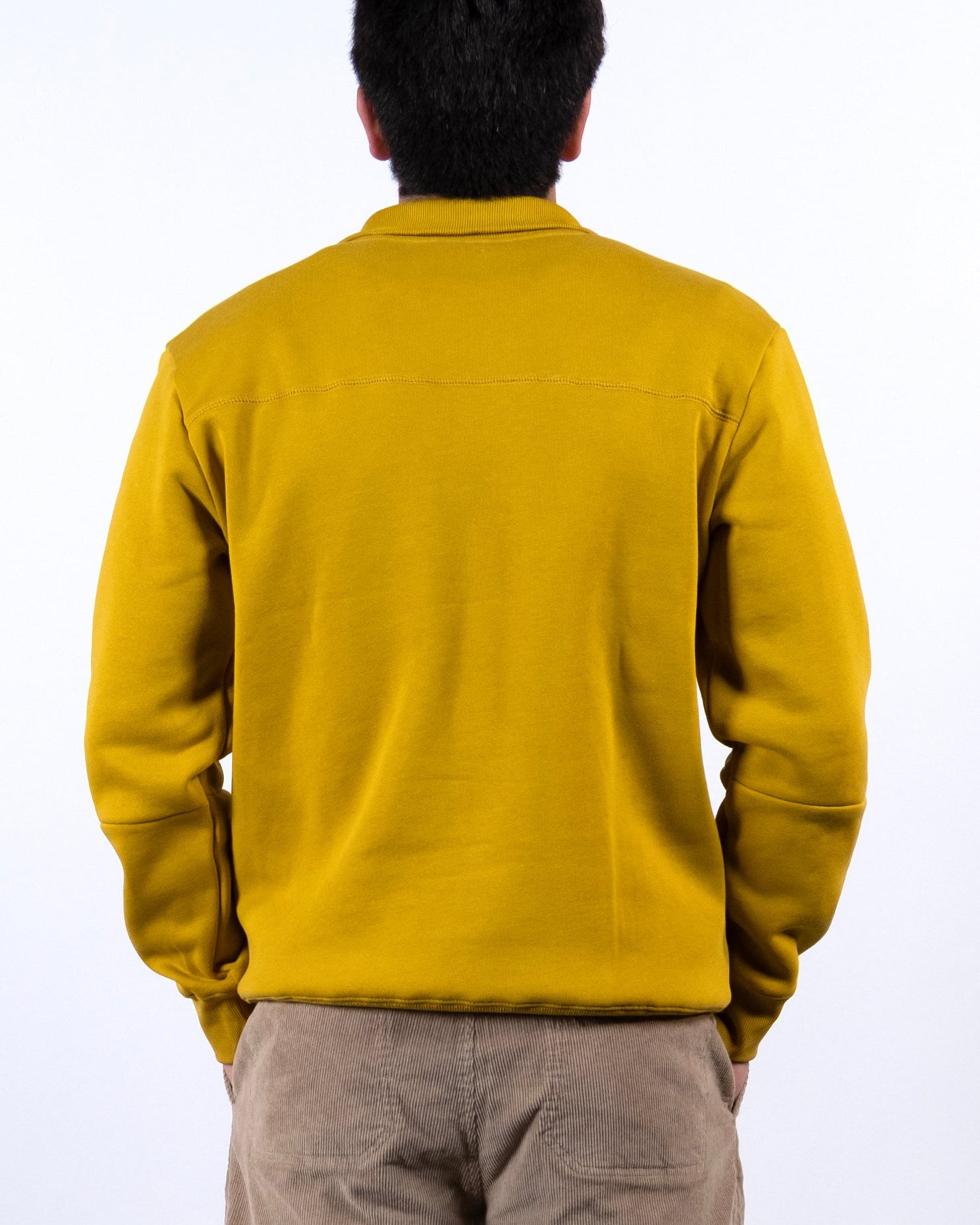 A person with short dark hair faces away from the camera, wearing the mustard yellow BEARDEDGOAT NEW — VOID™ Half Zip and beige shorts against a plain white background.