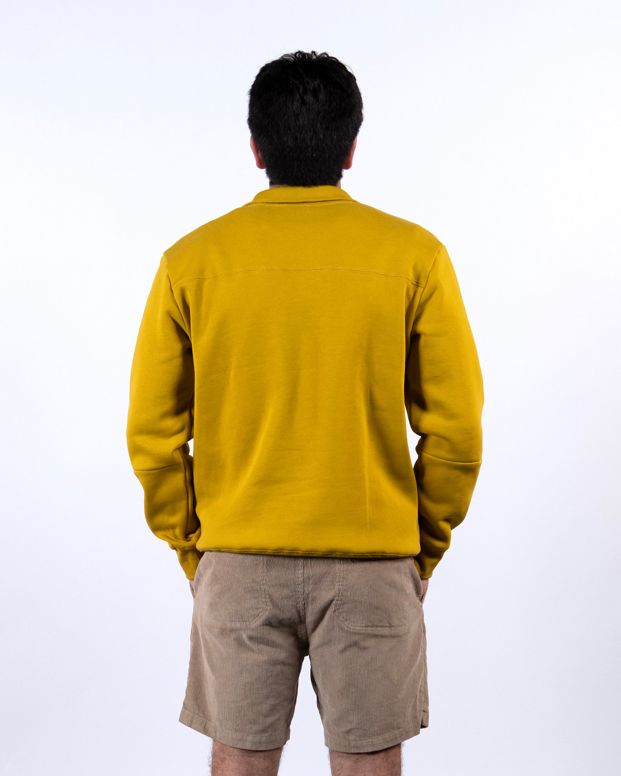 A person with short dark hair faces away from the camera, wearing the mustard yellow BEARDEDGOAT NEW — VOID™ Half Zip and beige shorts against a plain white background.