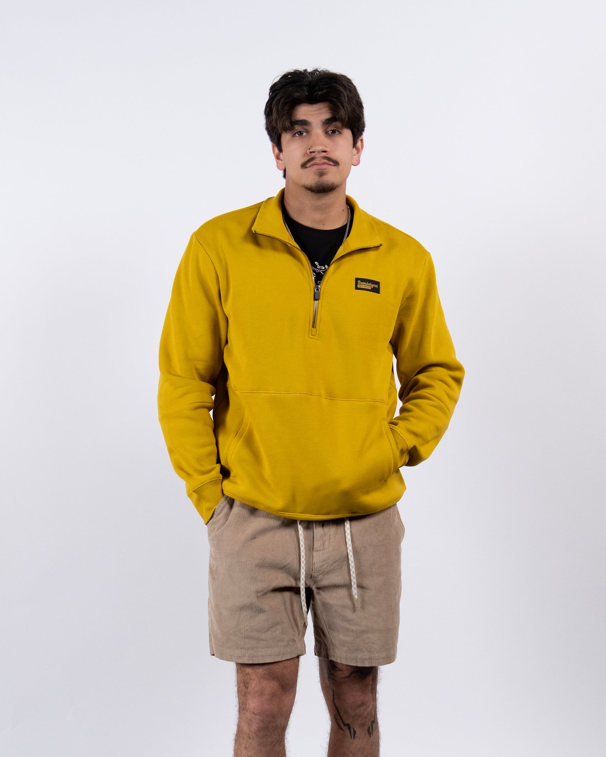 A young man with short dark hair stands against a plain white background, wearing the BEARDEDGOAT NEW — VOID™ Half Zip in mustard yellow over a black t-shirt and beige drawstring shorts, sporting a neutral expression.