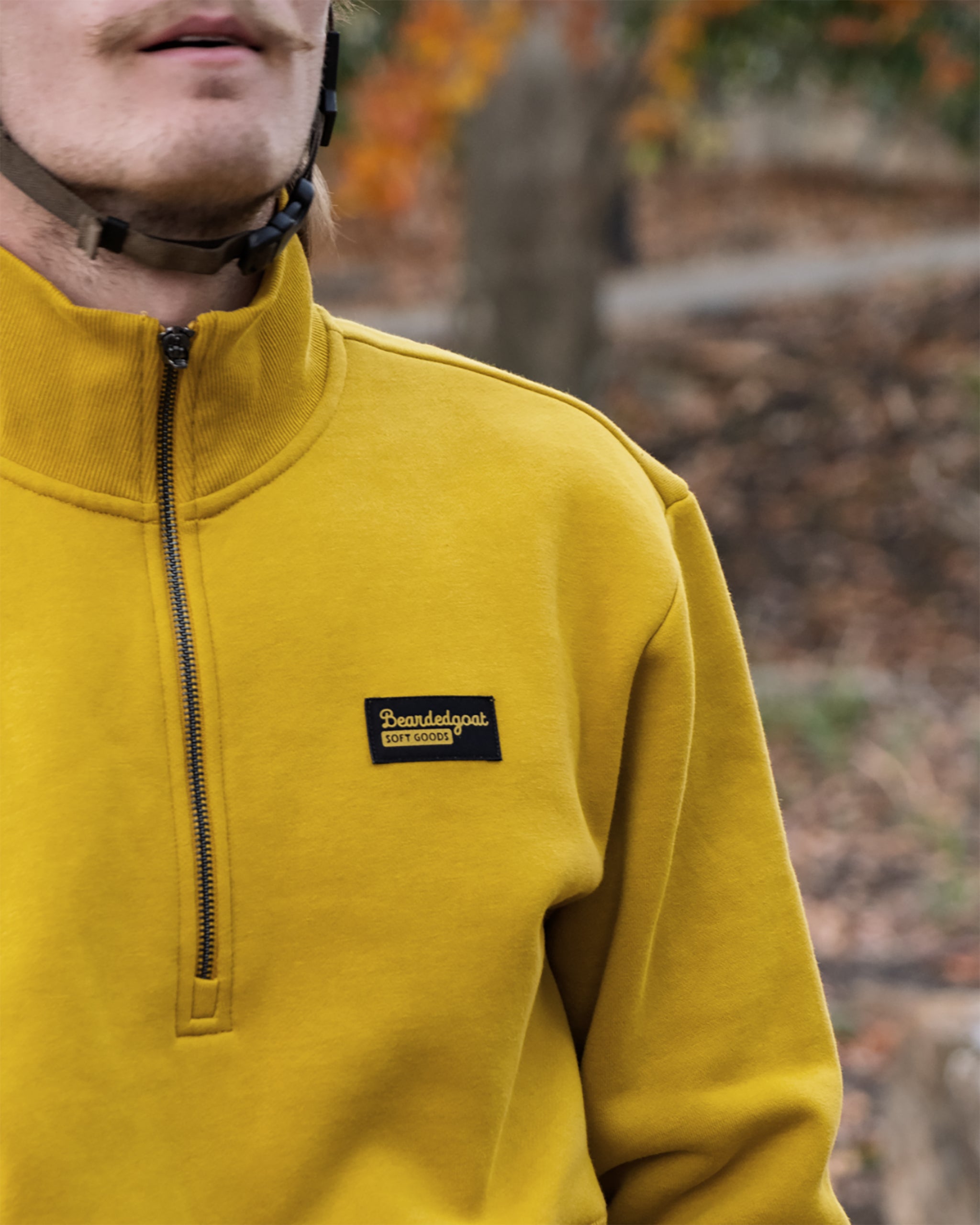A person wears the BEARDEDGOAT NEW — VOID™ Half Zip in mustard yellow premium cotton with a 100% local Soundiegoat patch and a helmet, standing outdoors with blurred trees in the background.