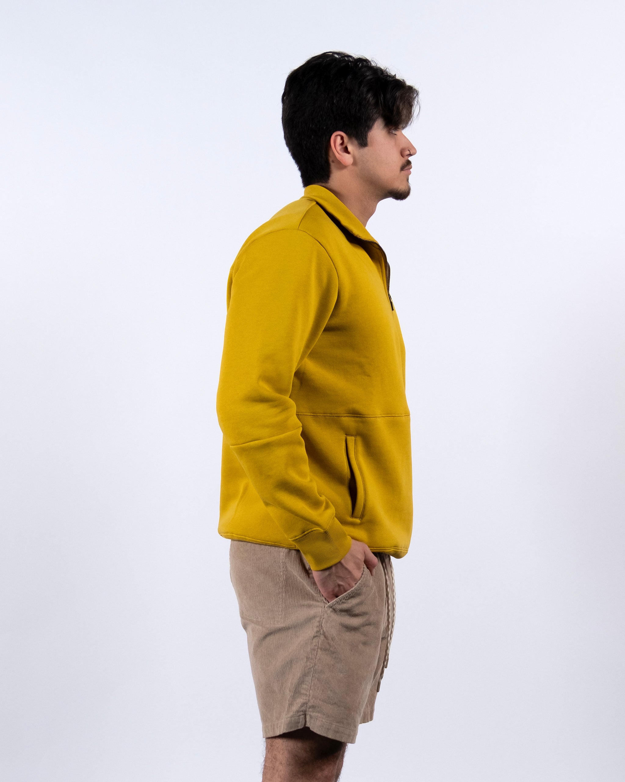 A person with dark hair, wearing the BEARDEDGOAT NEW — VOID™ Half Zip in yellow and beige shorts, stands in profile against a plain white background with one hand in their pocket.