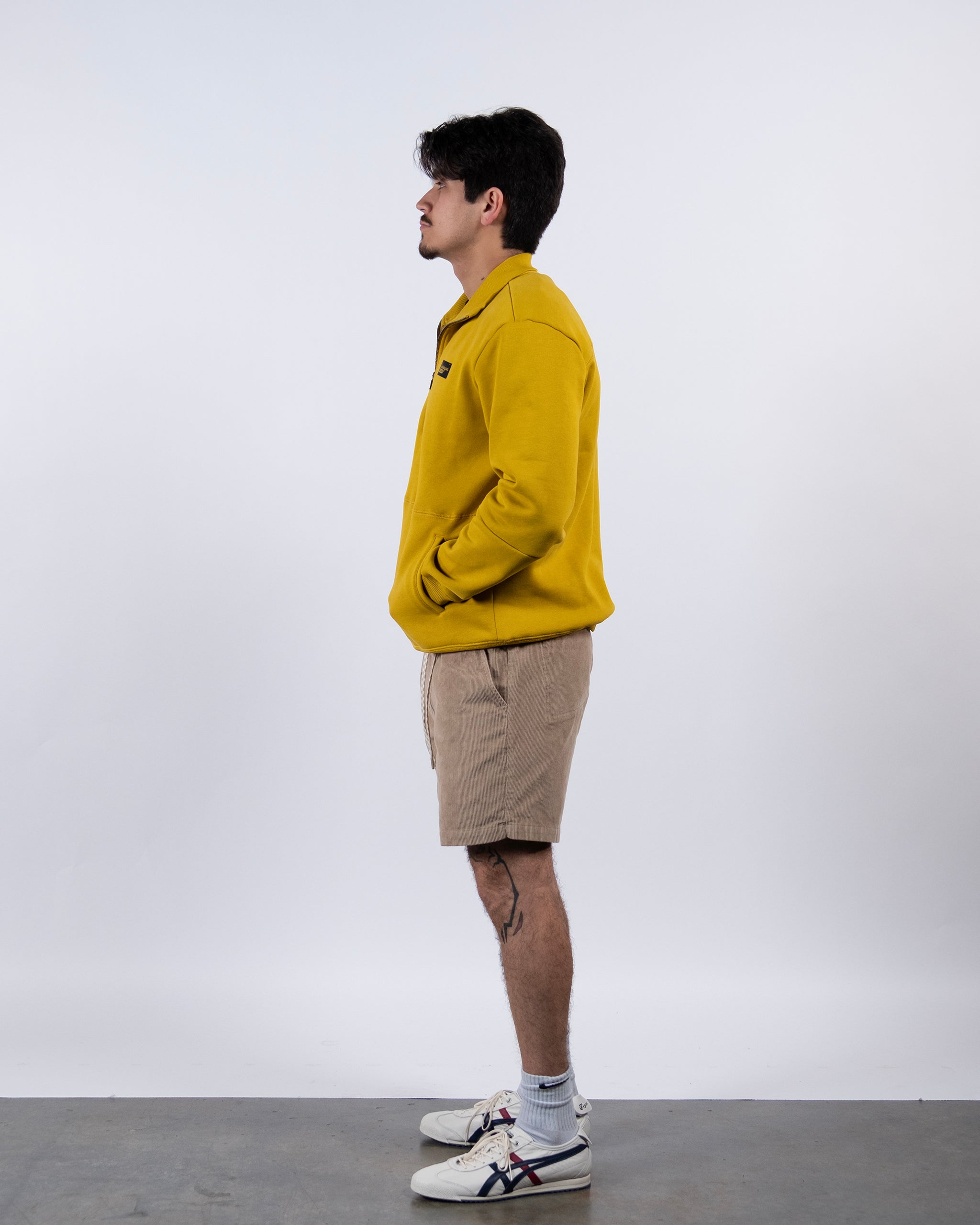 A young man stands in profile against a plain white background, wearing the BEARDEDGOAT NEW — VOID™ Half Zip, beige shorts, white socks, and white sneakers with dark accents. His hands are in his pockets and he looks forward.