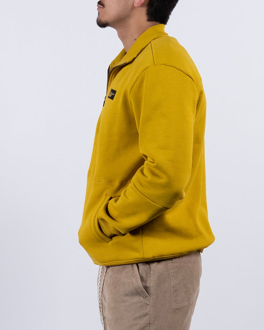 A young man stands in profile against a plain white background, wearing the BEARDEDGOAT NEW — VOID™ Half Zip, beige shorts, white socks, and white sneakers with dark accents. His hands are in his pockets and he looks forward.