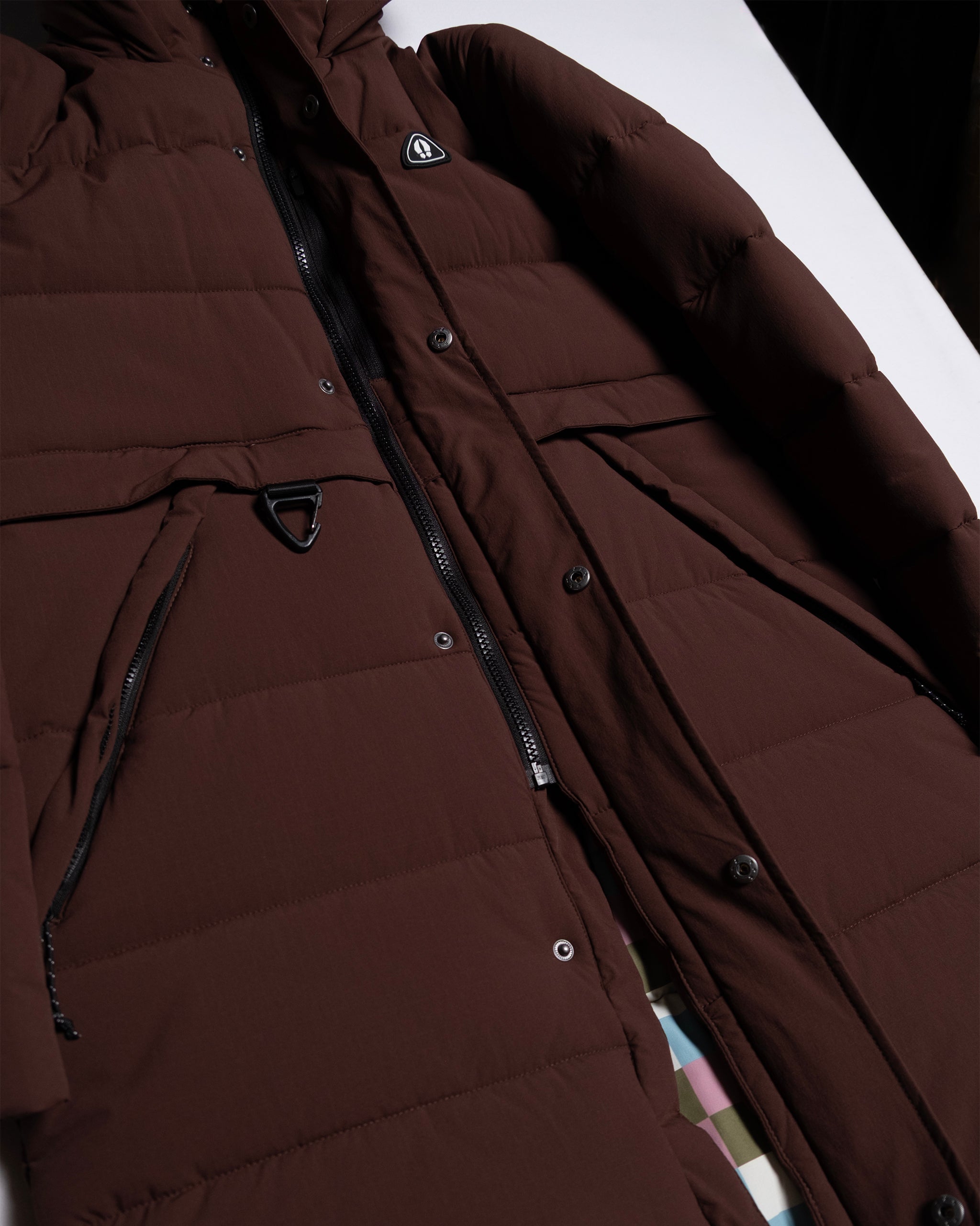 A close-up of the limited edition beardedgoat NEW — MERIDIAN™ PARKA, a brown synthetic down jacket with black zippers, snap buttons, geometric metal details, quilted design, and flap pockets, laid flat on a white surface.