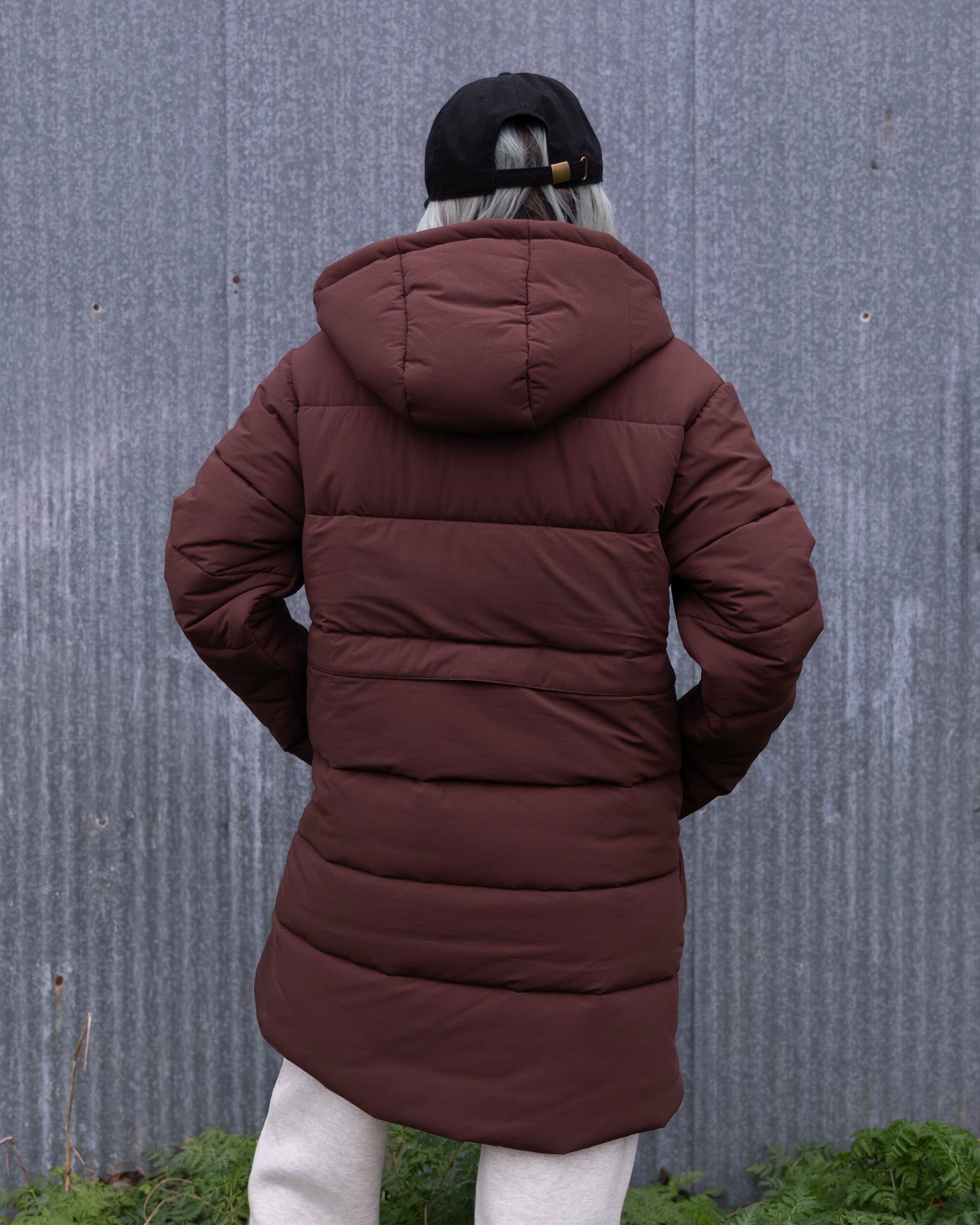 A person with gray hair, black cap, limited edition brown beardedgoat NEW — MERIDIAN™ PARKA with hood, and light pants stands outdoors facing a corrugated metal wall bordered by green plants.