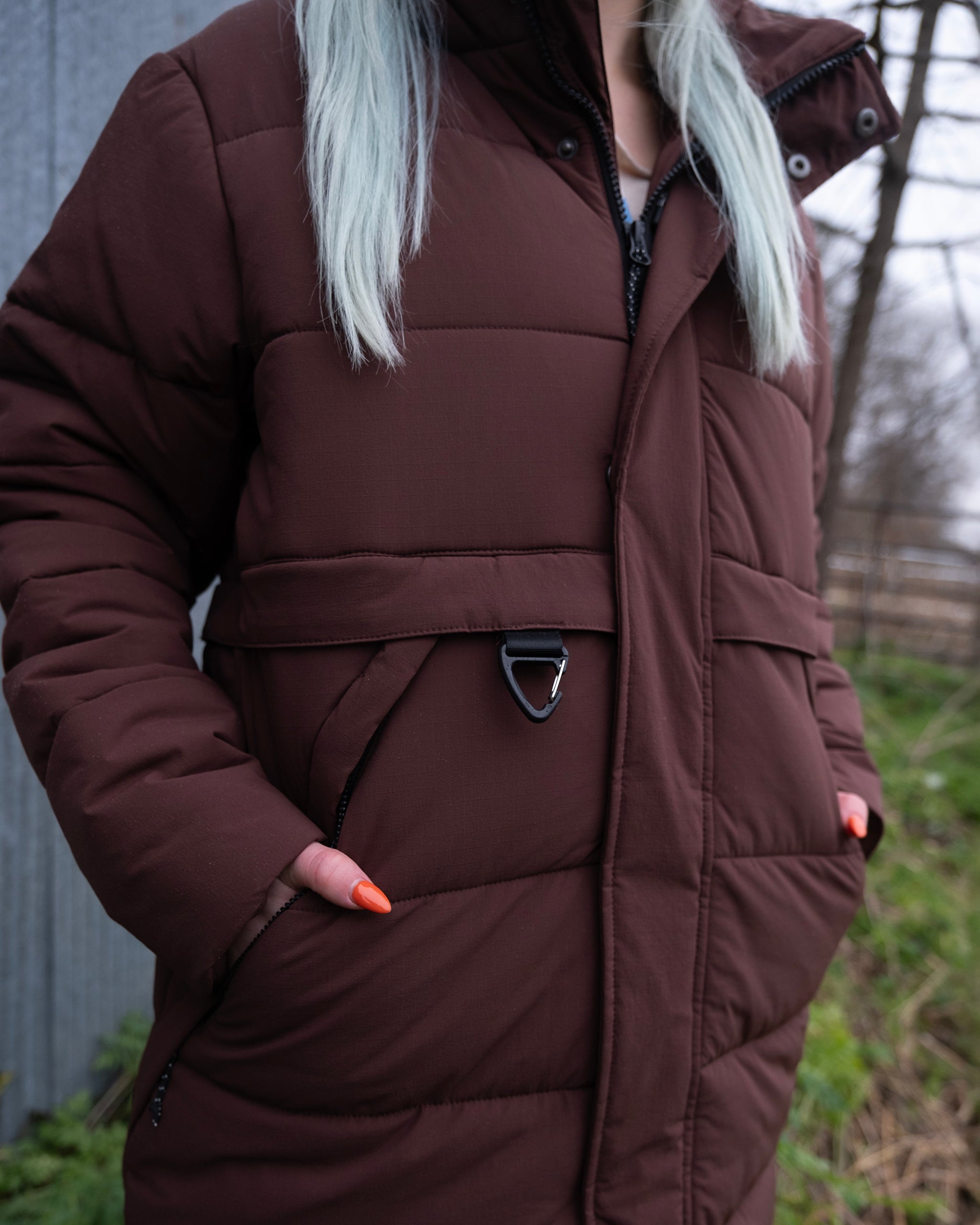 A person with long, light-colored hair wears the limited edition beardedgoat NEW — MERIDIAN™ PARKA outdoors, hands in pockets showing orange nails. Grass and a blurred fence are visible in the background.