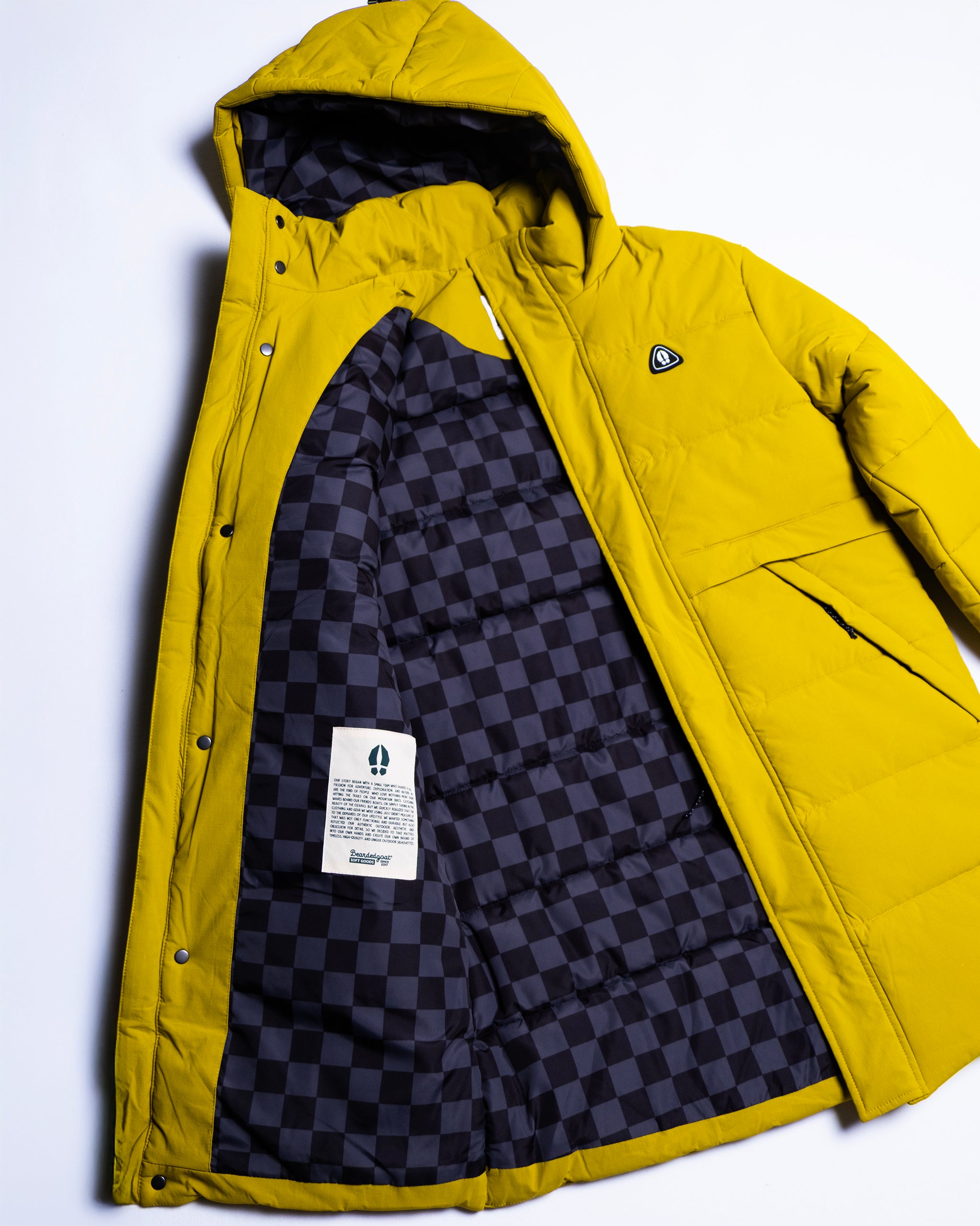 The NEW — MERIDIAN™ PARKA by beardedgoat is a yellow hooded, limited edition synthetic down parka with snap buttons, a small chest logo, black and dark gray checkered lining, and a white inner tag.