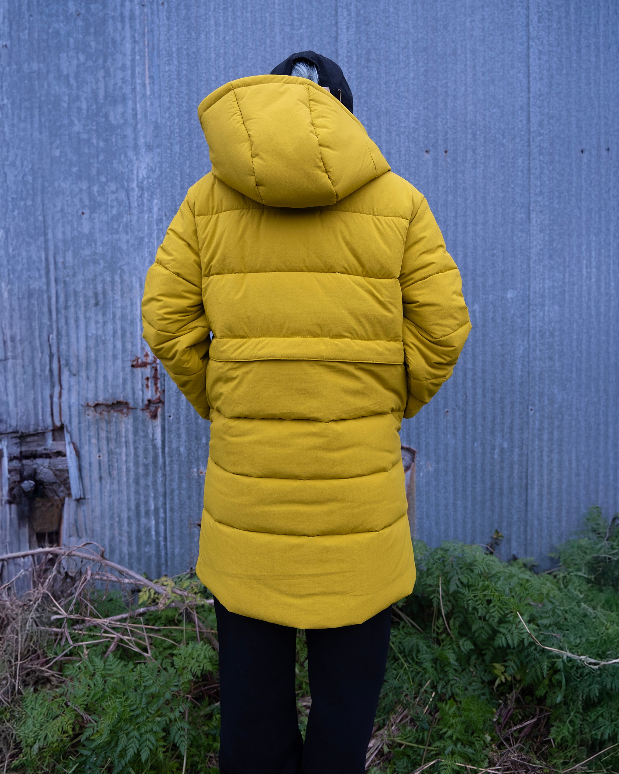 A person wearing the beardedgoat NEW — MERIDIAN™ PARKA, a long yellow limited edition synthetic down parka with a hood, stands facing away from the camera in front of a weathered metal wall with greenery at their feet.