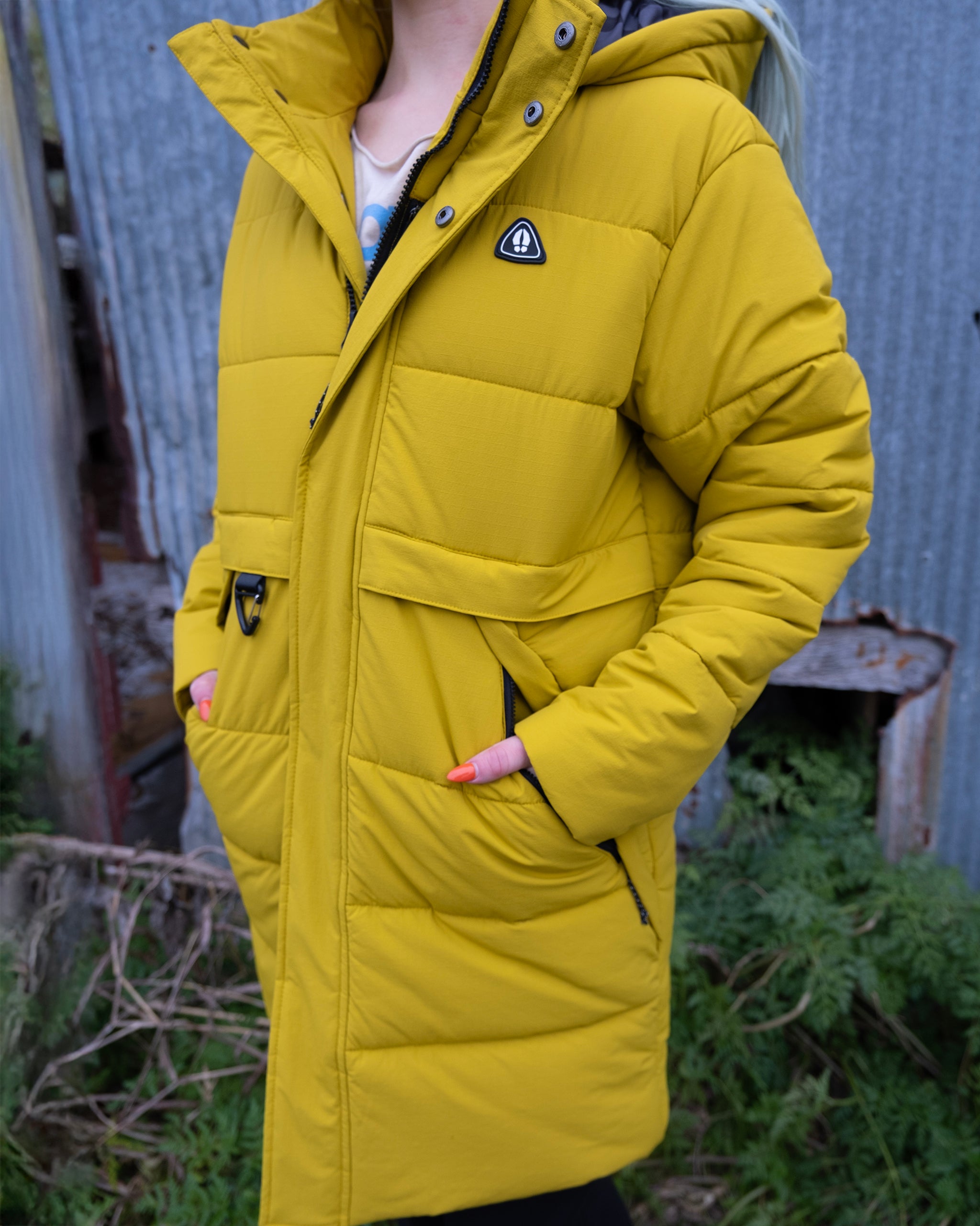 A person outdoors wears the limited edition beardedgoat NEW — MERIDIAN™ PARKA in bright yellow, featuring synthetic down, quilted stitching, and a chest badge, with greenery and a corrugated metal wall behind them.