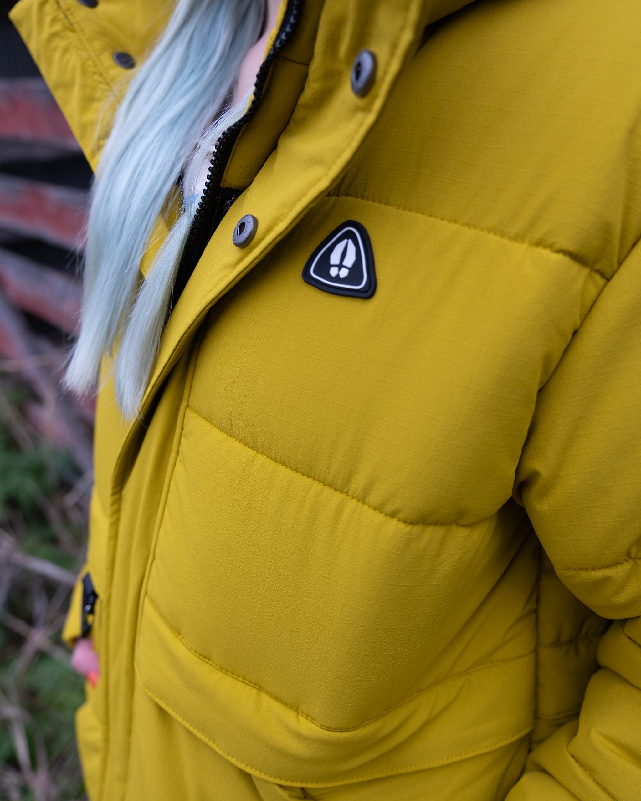 A person with long, light blue hair models the beardedgoat NEW — MERIDIAN™ PARKA, a bright yellow synthetic down jacket featuring a black and white triangular patch. The photo is shot from the shoulders down with a blurred wall and grass in the background.