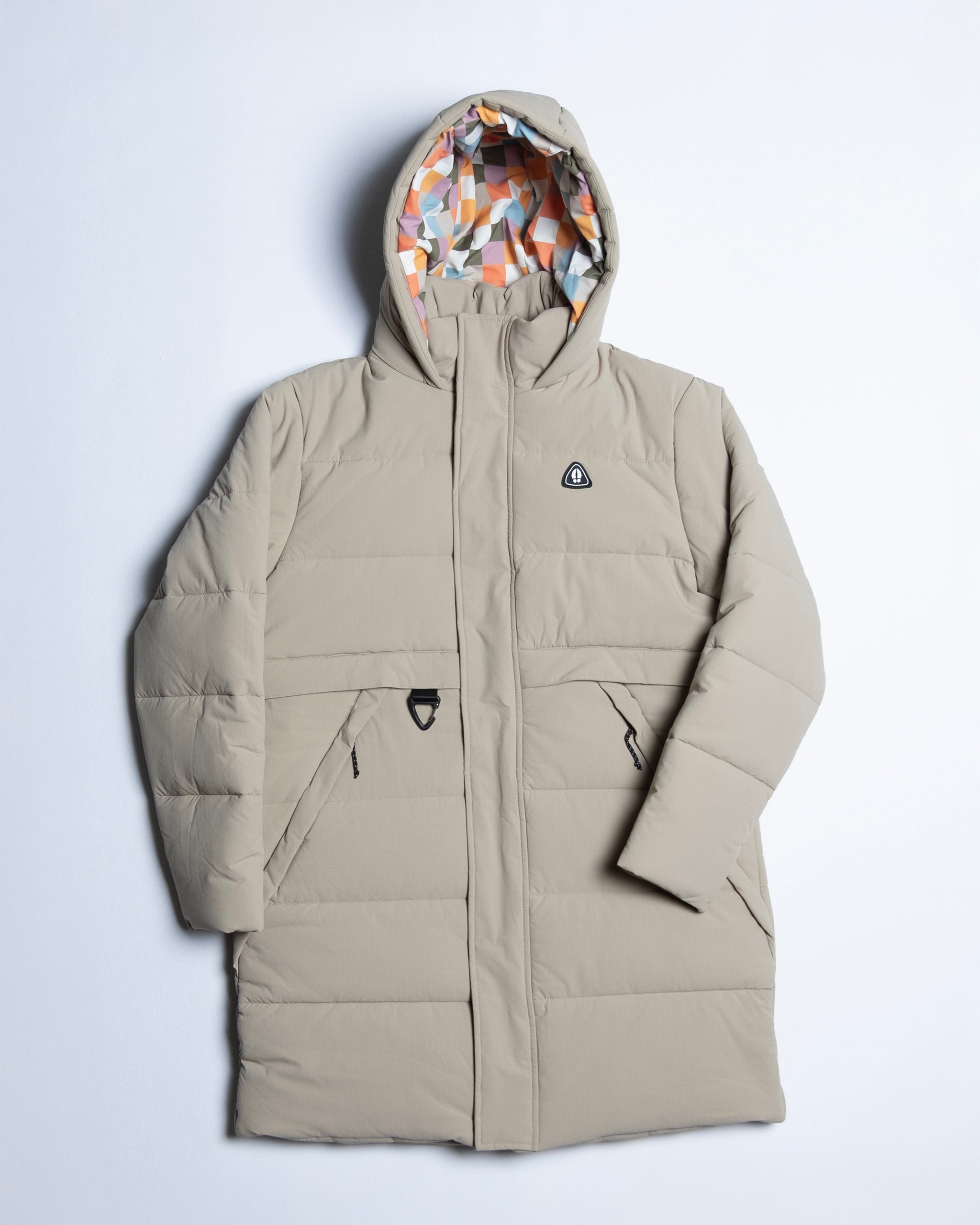 The NEW — MERIDIAN™ PARKA by beardedgoat is a long, beige limited edition parka with synthetic down, zippered pockets, a black chest patch, and a hood lined with an orange, gray, and white geometric pattern.
