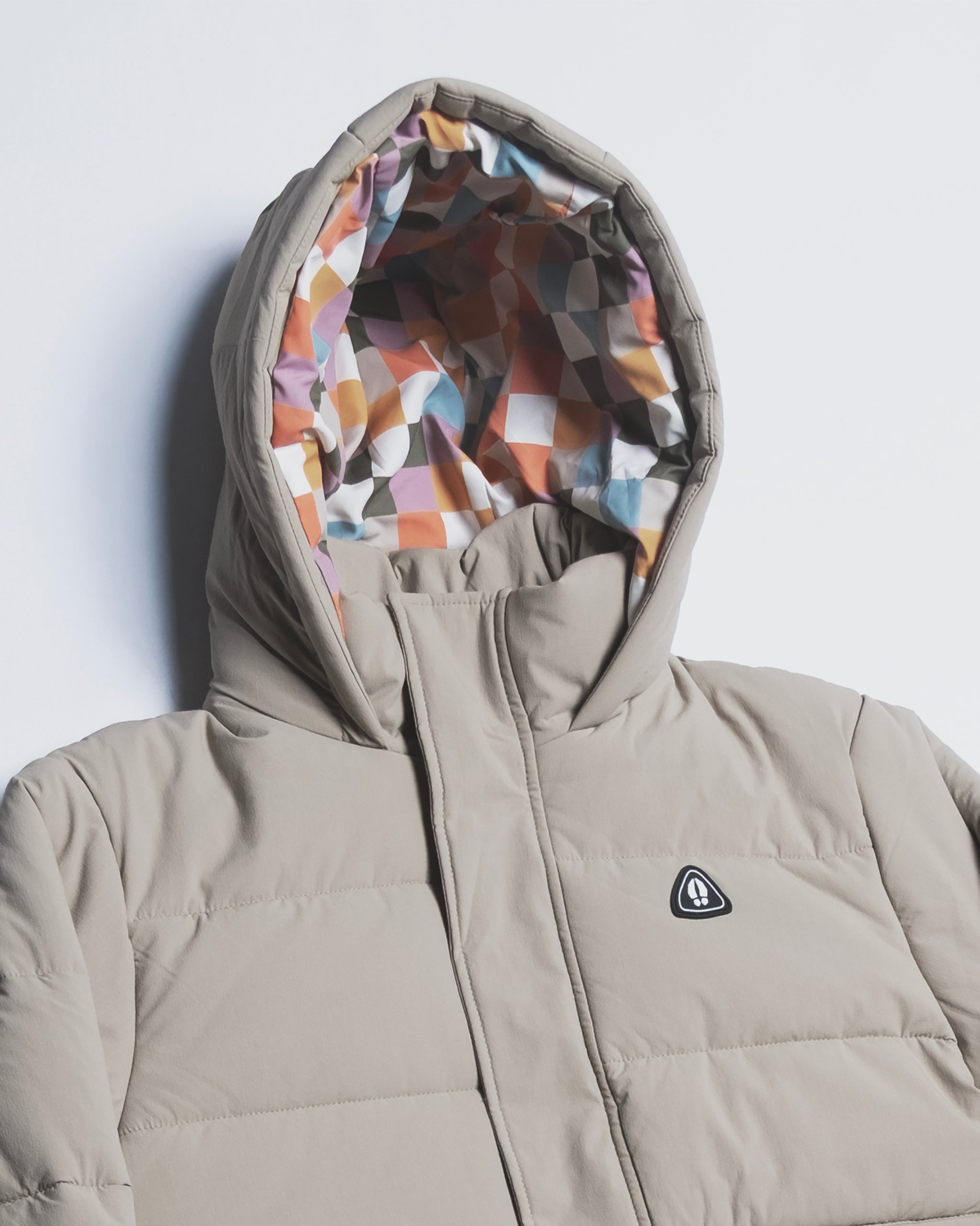 The NEW — MERIDIAN™ PARKA by beardedgoat is a beige synthetic down hooded parka with a colorful geometric lining and a black and white triangular chest logo, shown on a white background. This limited edition blends style with warmth.