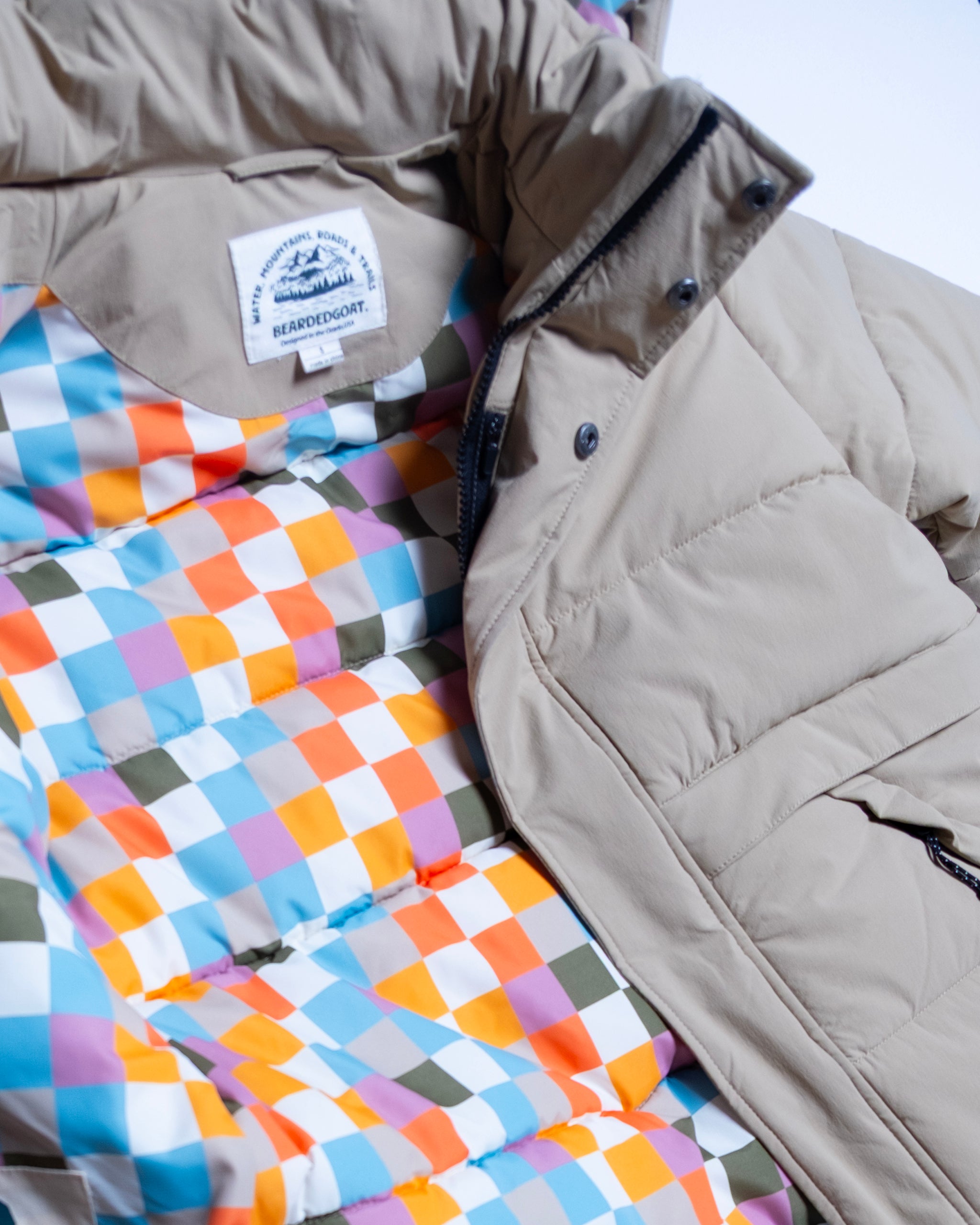 The NEW — MERIDIAN™ PARKA by beardedgoat is a beige synthetic down parka featuring a colorful checkered blue, orange, white, green, and pink lining. It's shown partially unzipped to reveal an inner label beneath the collar.