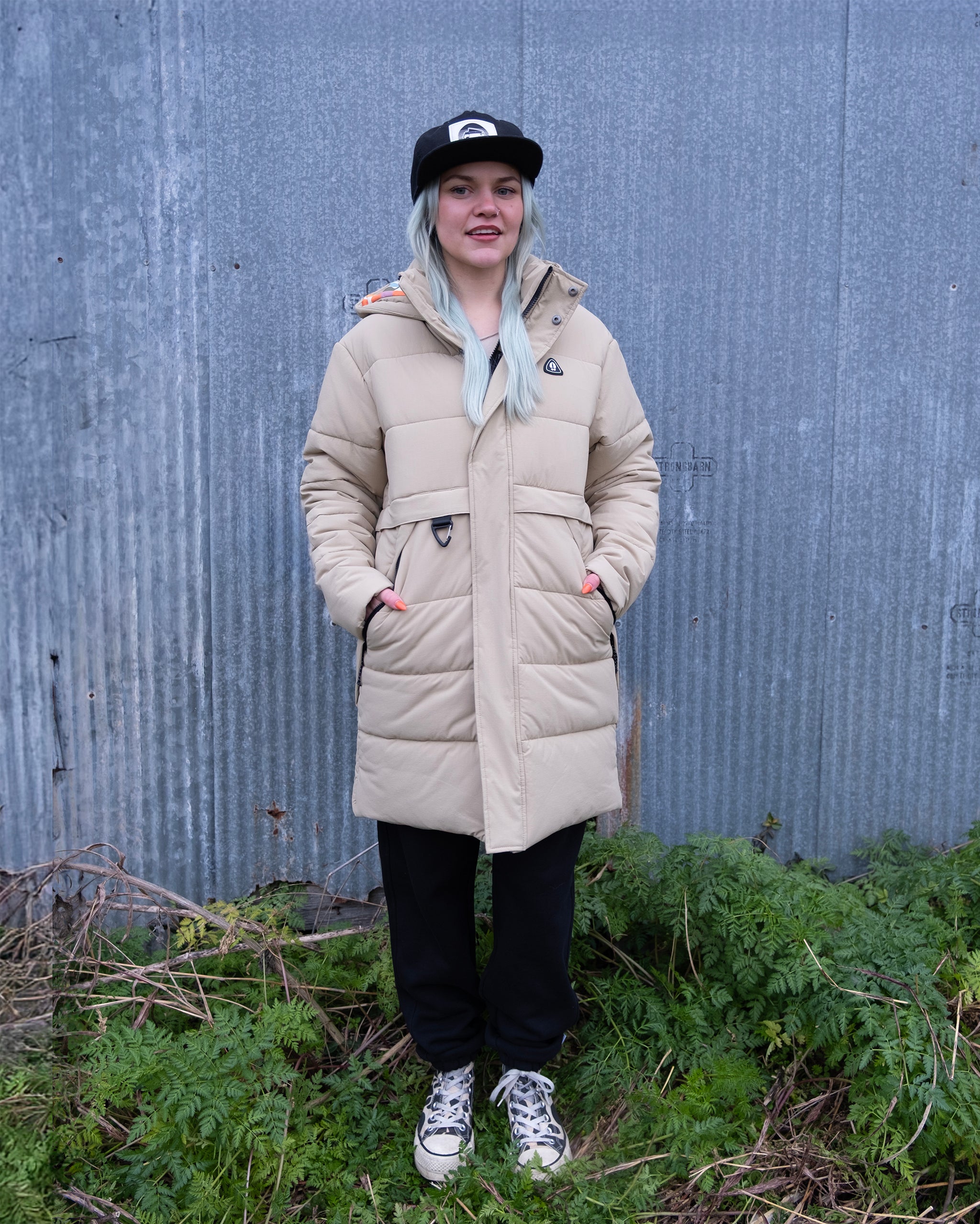 A person with light blue hair, dressed in the NEW — MERIDIAN™ PARKA by beardedgoat in Terra Sienna, black pants, patterned sneakers, and a black cap stands on grass before a corrugated metal wall.