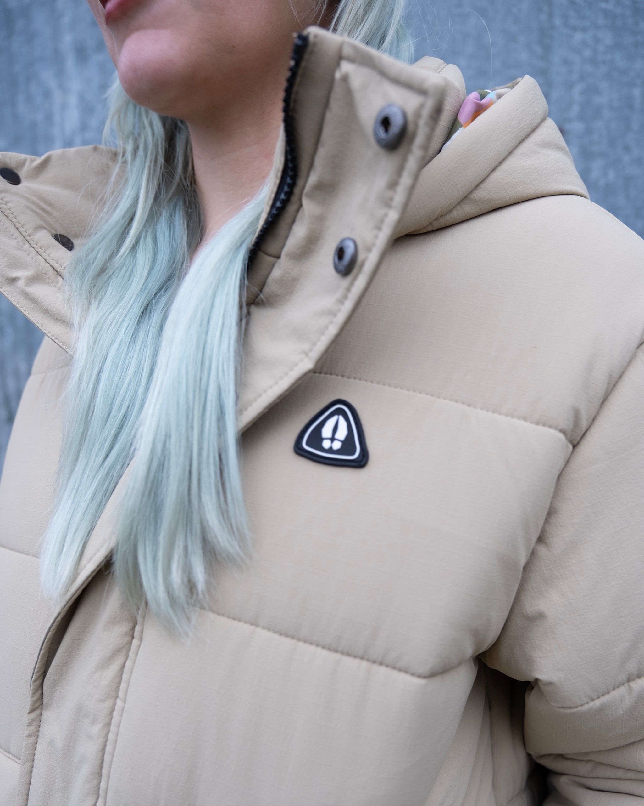 A person with long, light blue hair wears the limited edition beardedgoat NEW — MERIDIAN™ PARKA featuring a black-and-white triangular chest patch, standing against a gray textured background.