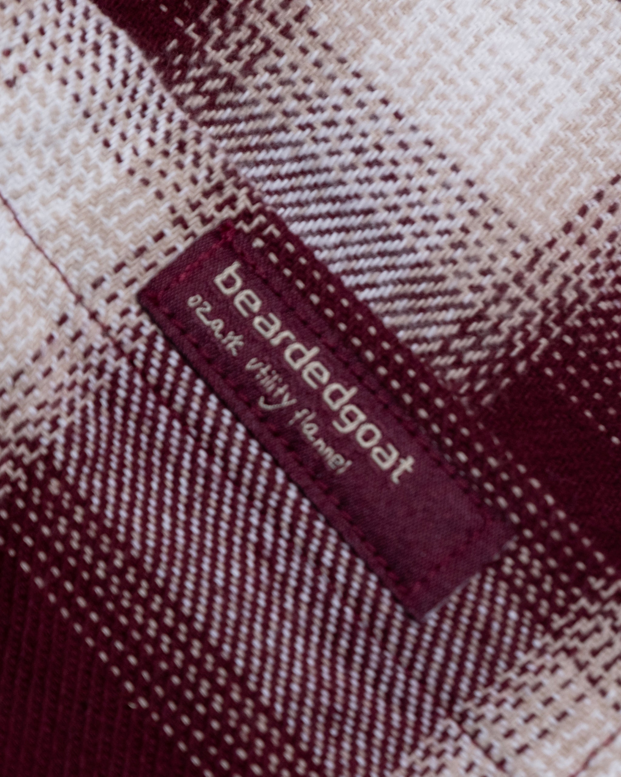 A close-up of the NEW--Ozark Utility Flannel™ by beardedgoat, featuring maroon, cream, and brown plaid heavyweight cotton fabric with a maroon tag that reads “beardedgoat” and the slogan “break with the herd” in white.