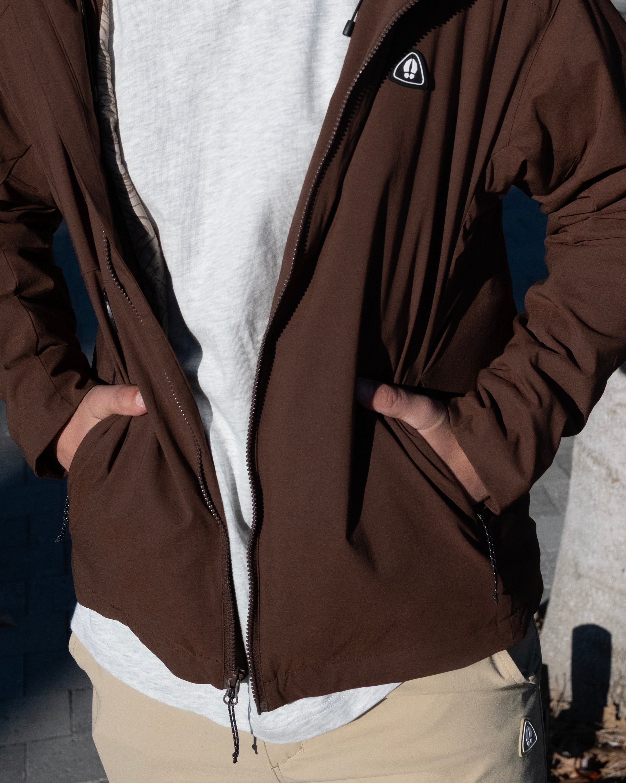 A person wears a beardedgoat NEW — OSLO Rover™ Performance All-Season Jacket in brown over a light gray shirt and khaki pants, standing with hands in the jacket pockets. The photo is cropped to show only the torso.