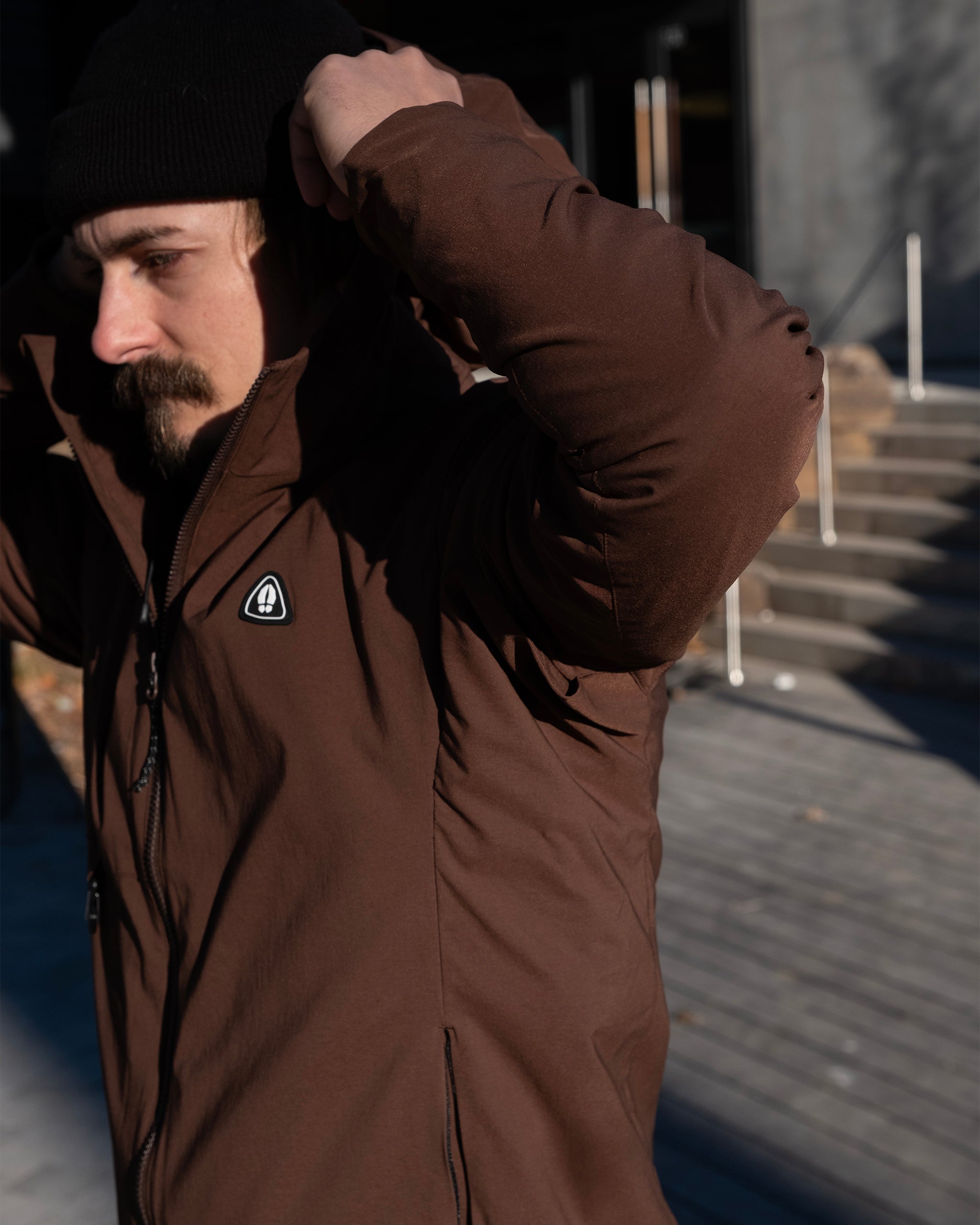 A man in a Chickory beardedgoat NEW — OSLO Rover™ Performance All-Season Jacket and black beanie adjusts his hood outside on a sunny day, with steps and a partially shadowed building in the background.