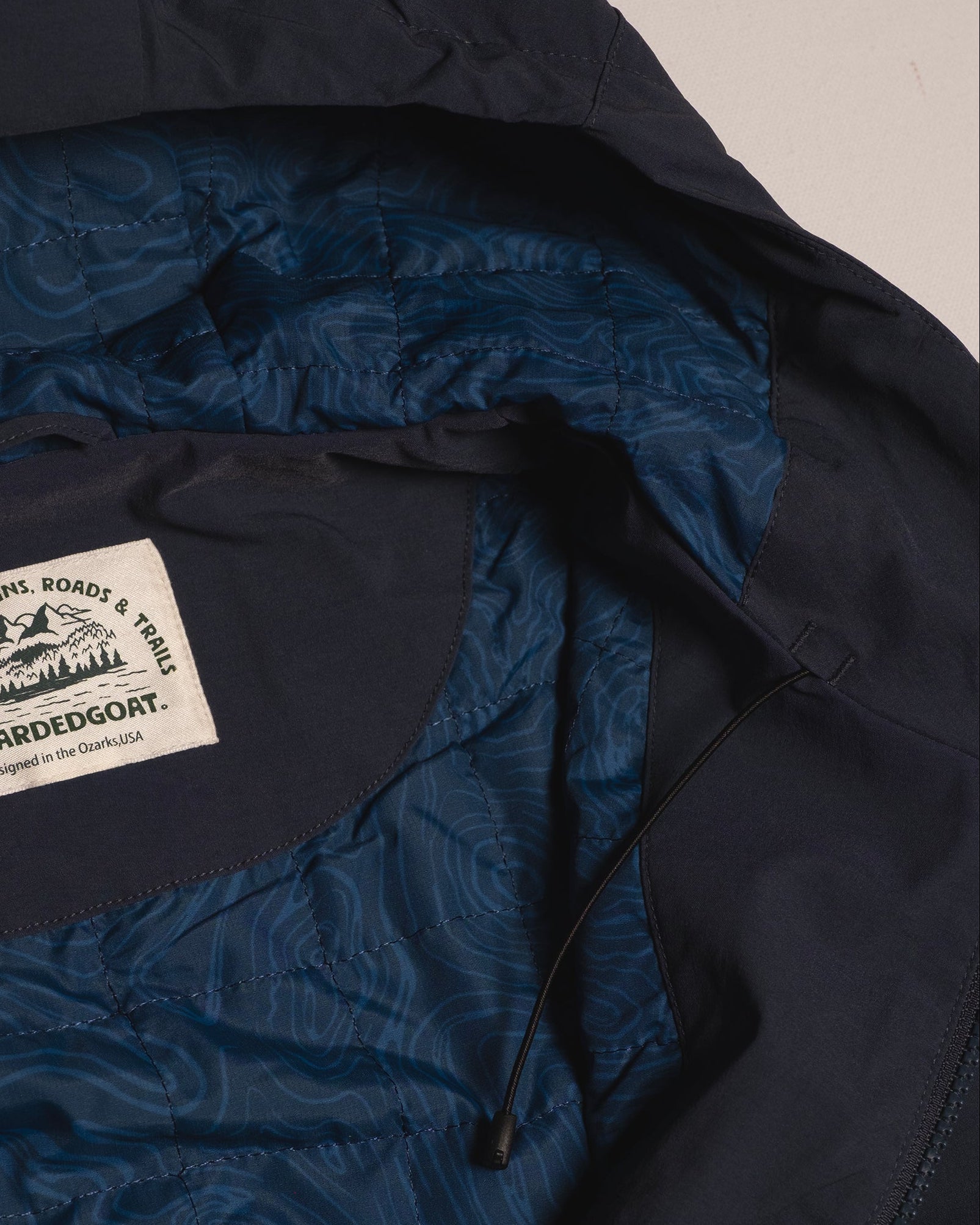 Close-up of the NEW — OSLO Rover™ Performance All-Season Jacket by beardedgoat, a navy all-season jacket with quilted patterned lining, mountain-logo "beardedgoat" label, and a black drawstring for added functionality.