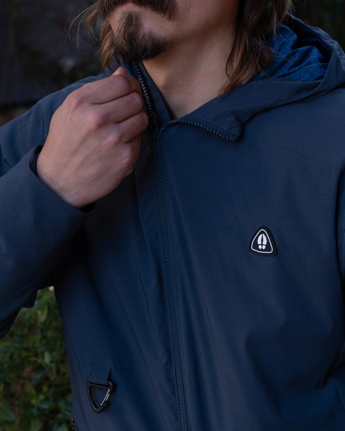 A person with long hair and a mustache zips up the beardedgoat NEW — OSLO Rover™ Performance All-Season Jacket, showcasing its dark blue color and small chest logo, with only their lower face and upper body visible.