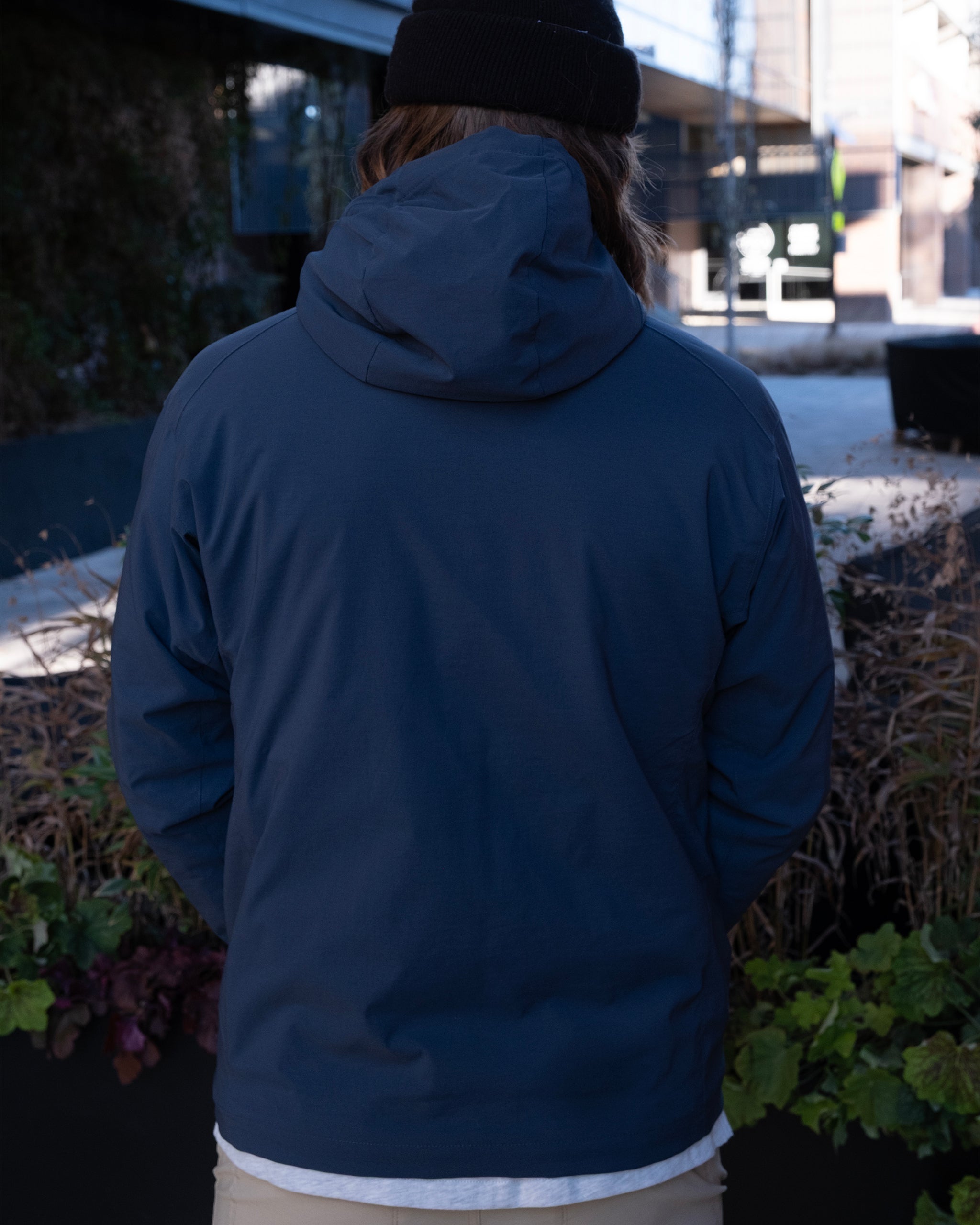 A person in a dark blue beardedgoat NEW — OSLO Rover™ Performance All-Season Jacket and black beanie stands outdoors with their back to the camera, surrounded by plants and city buildings.