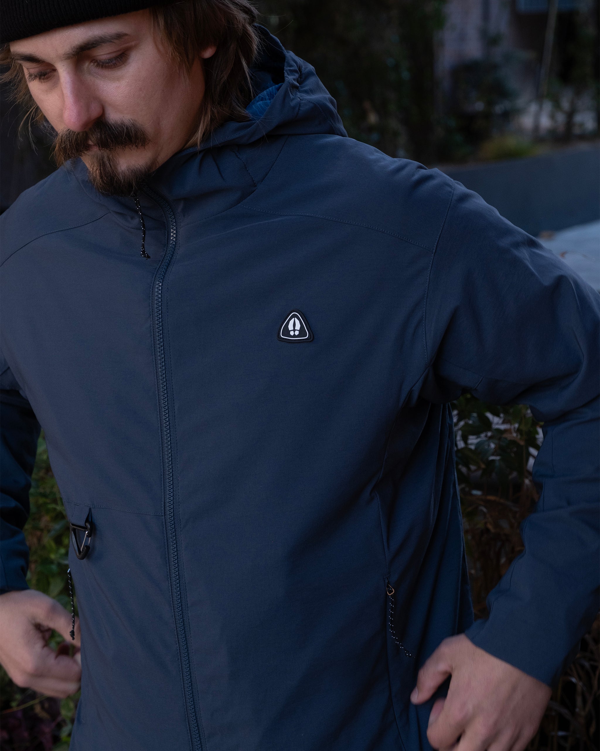 A man with a mustache in a black beanie and the beardedgoat NEW — OSLO Rover™ Performance All-Season Jacket in Mid Night Navy stands outdoors, looking down with his hands near his pockets. Trees and greenery are in the background.