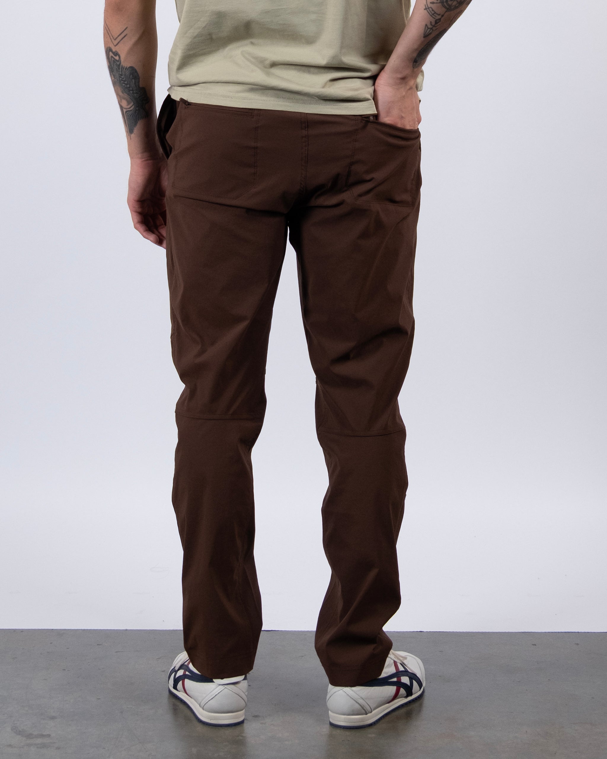 A person stands on a gray floor facing away, wearing BEARDEDGOAT's NEW — ROVER™ Pant in brown, a beige shirt, and white sneakers with dark stripes; tattoos are visible on their left arm.