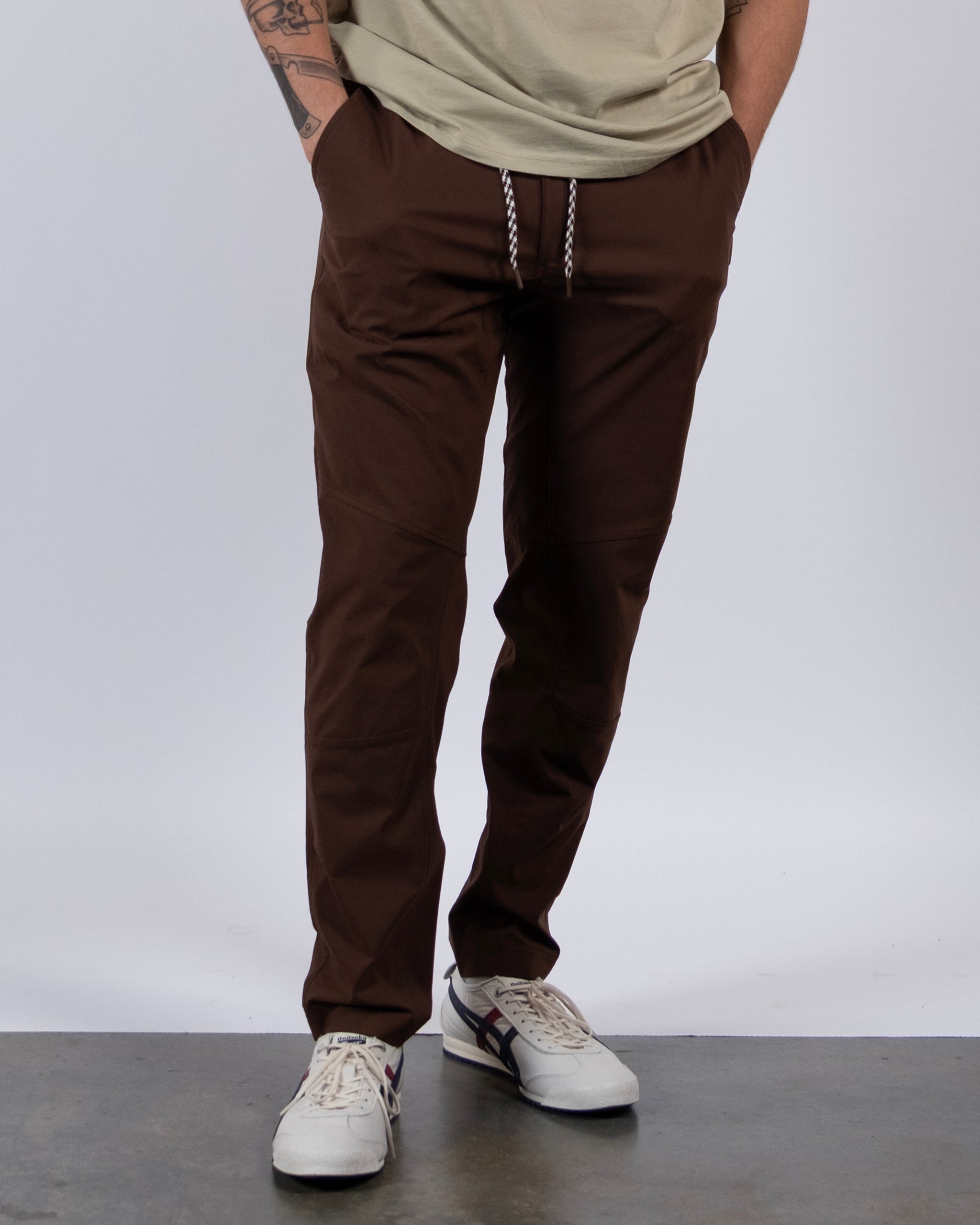 A person wearing BEARDEDGOAT’s NEW — ROVER™ Pant stands on a gray floor with hands in pockets, paired with a light beige shirt and white sneakers with dark accents; only the lower body is visible.
