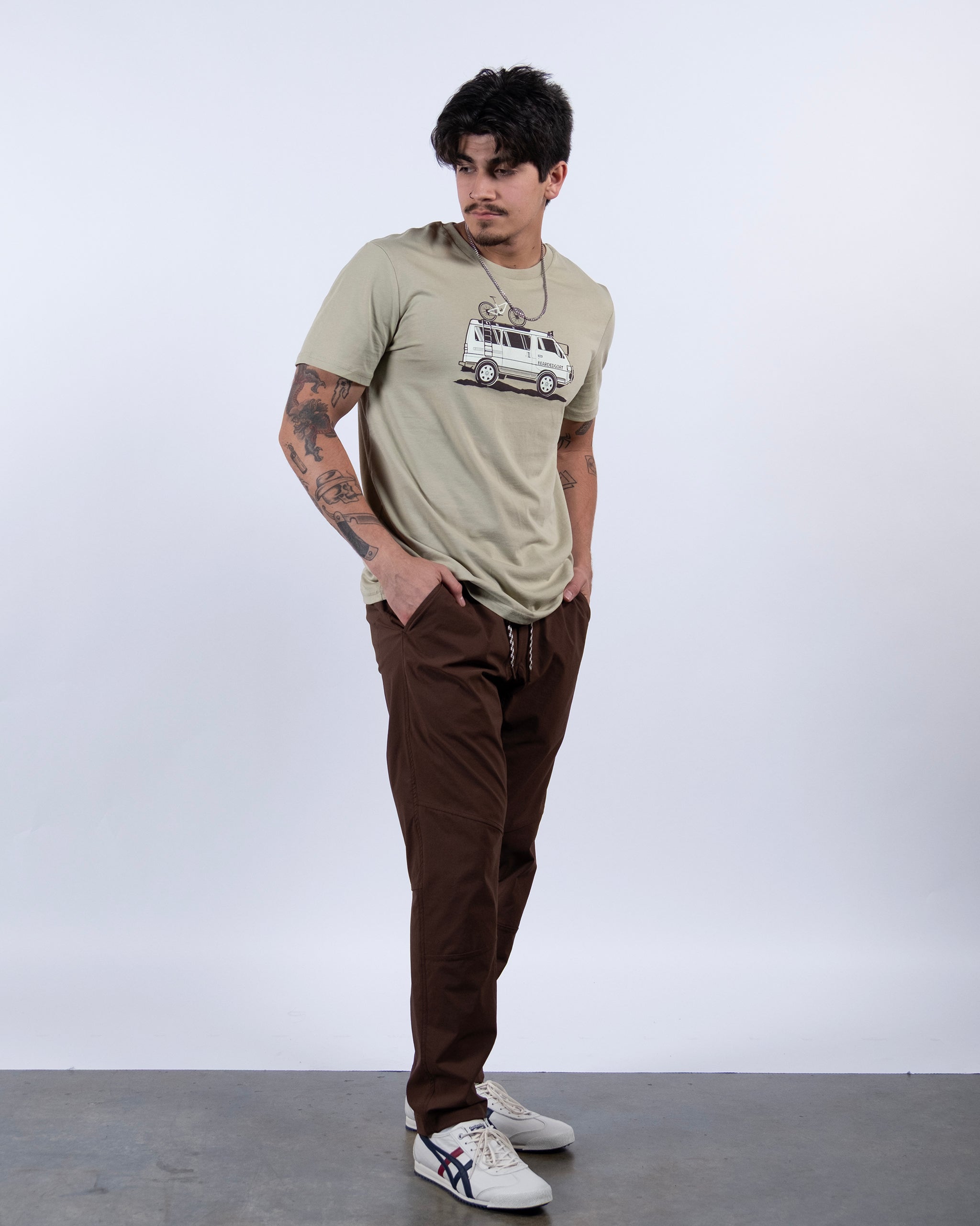A tattooed man stands with hands in pockets, wearing a beige graphic tee, BEARDEDGOAT NEW — ROVER™ Pant made from performance stretch nylon, and white sneakers. He looks to the side against a plain light background.