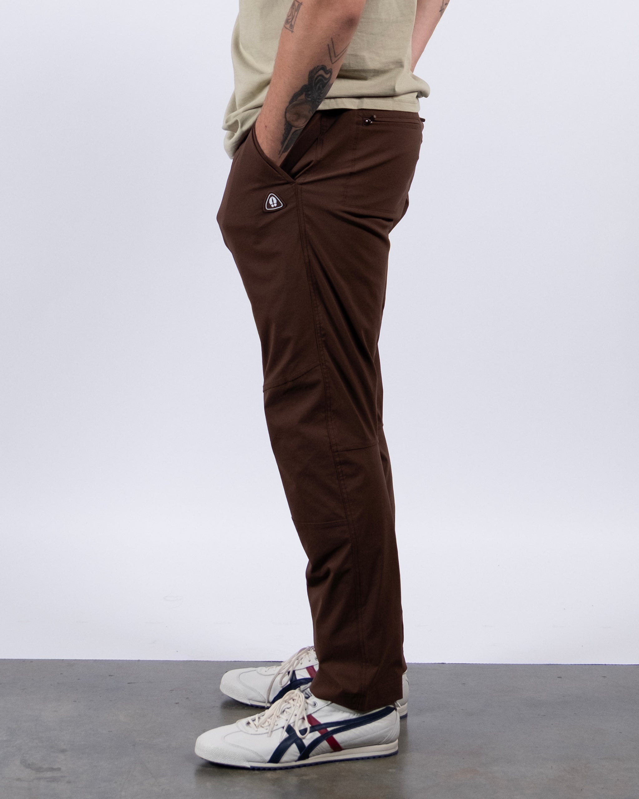 A person stands sideways in BEARDEDGOAT's NEW — ROVER™ Pant, made from brown performance stretch nylon, paired with a light shirt and white sneakers. Hands in pockets show a tattooed left forearm. The background is plain and light-colored.