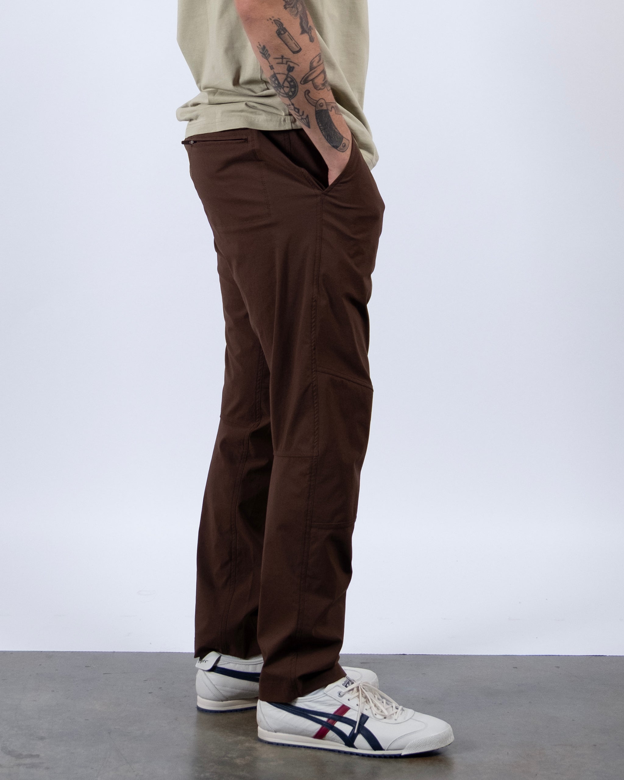 A person stands sideways with one hand in their pocket, showing arm tattoos. They wear a beige t-shirt, BEARDEDGOAT NEW — ROVER™ Pant, and white sneakers with blue and red accents. The background is plain white.