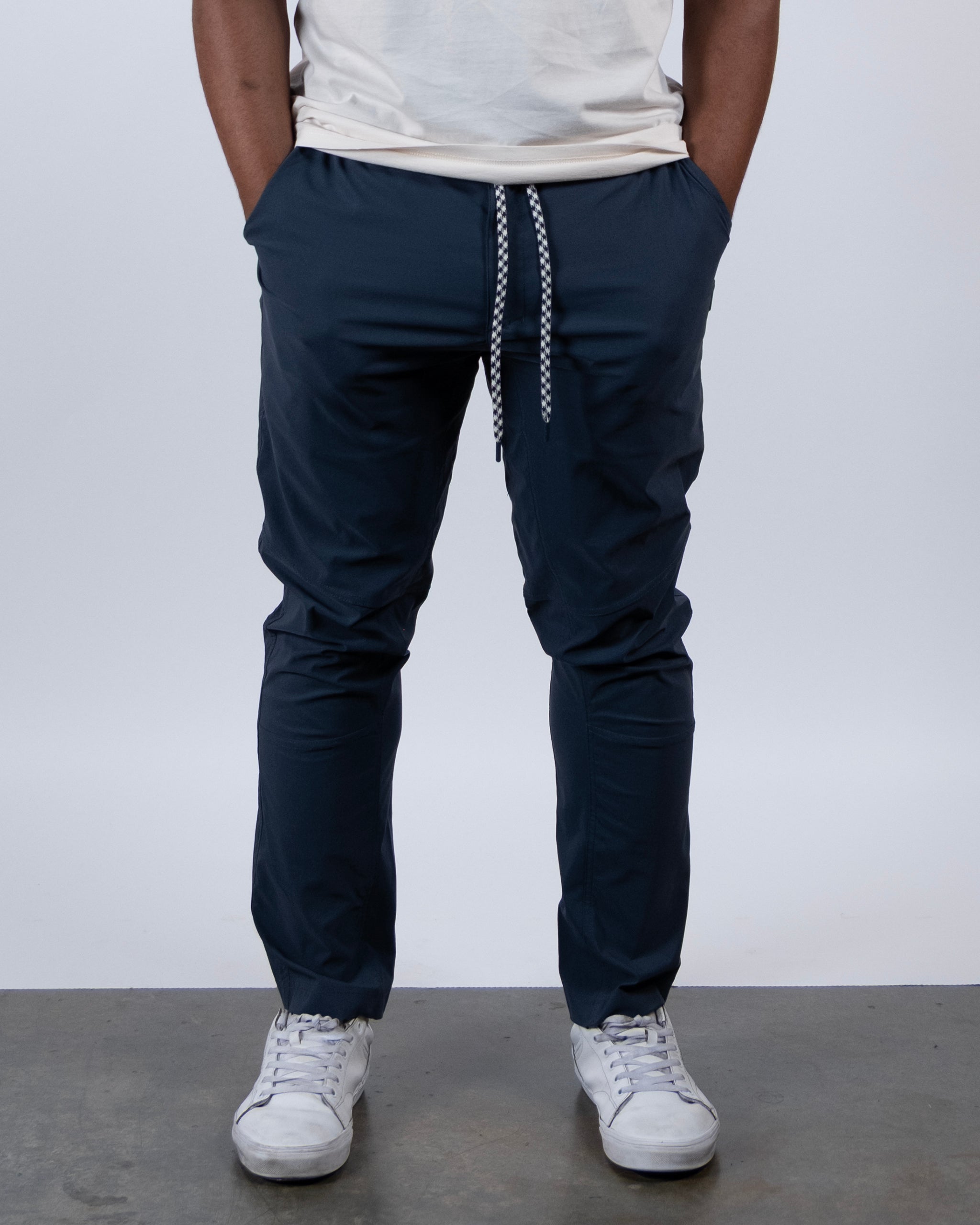 A person stands on a concrete floor with hands in pockets, wearing a white t-shirt, white sneakers, and BEARDEDGOAT NEW — ROVER™ Pant in Mid Night Navy. The image is cropped at the shoulders.