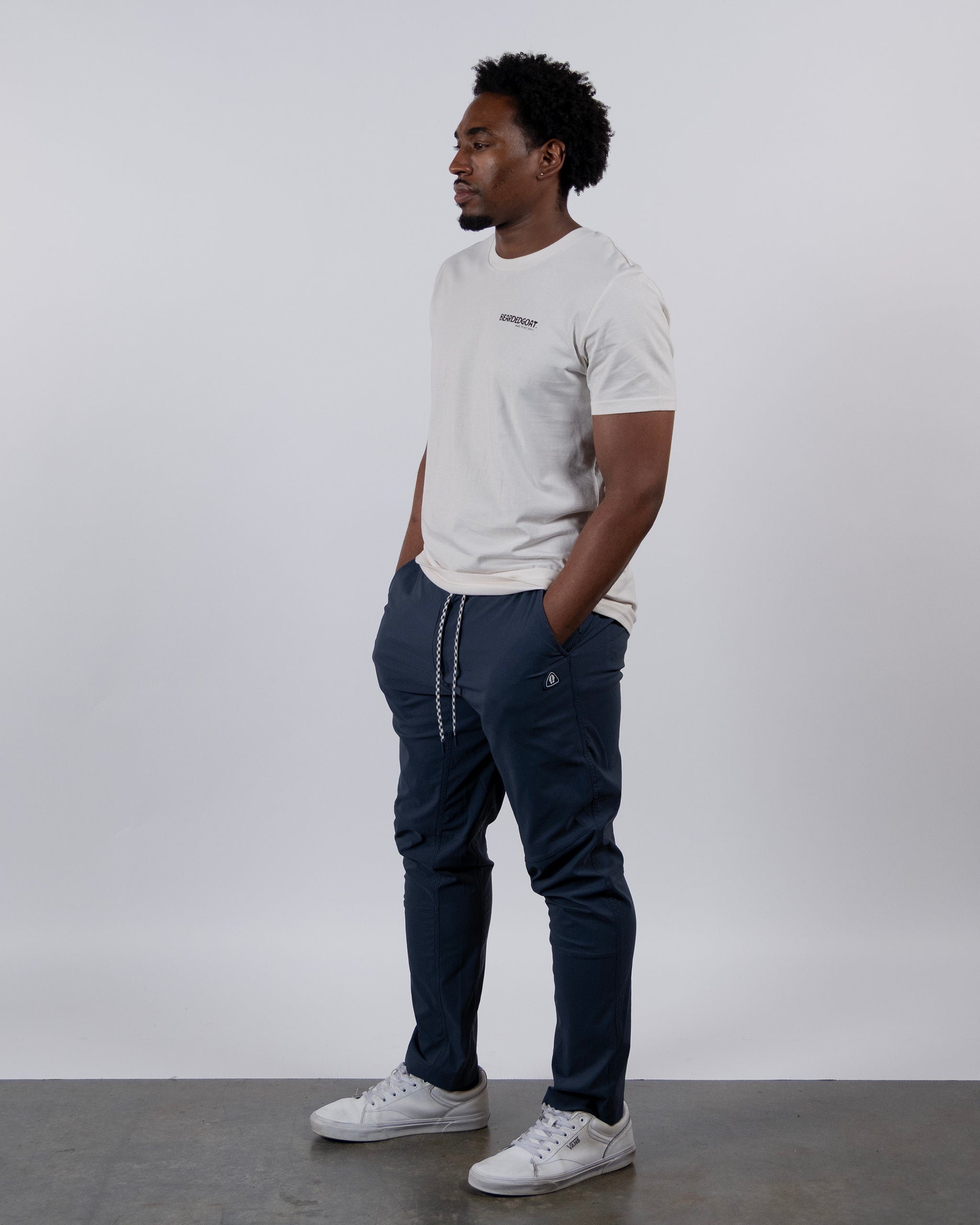 A man stands against a white background, wearing a white T-shirt, white sneakers, and BEARDEDGOAT’s NEW — ROVER™ Pant in dark blue with drawstrings. He has his hands in his pockets and looks to the side.