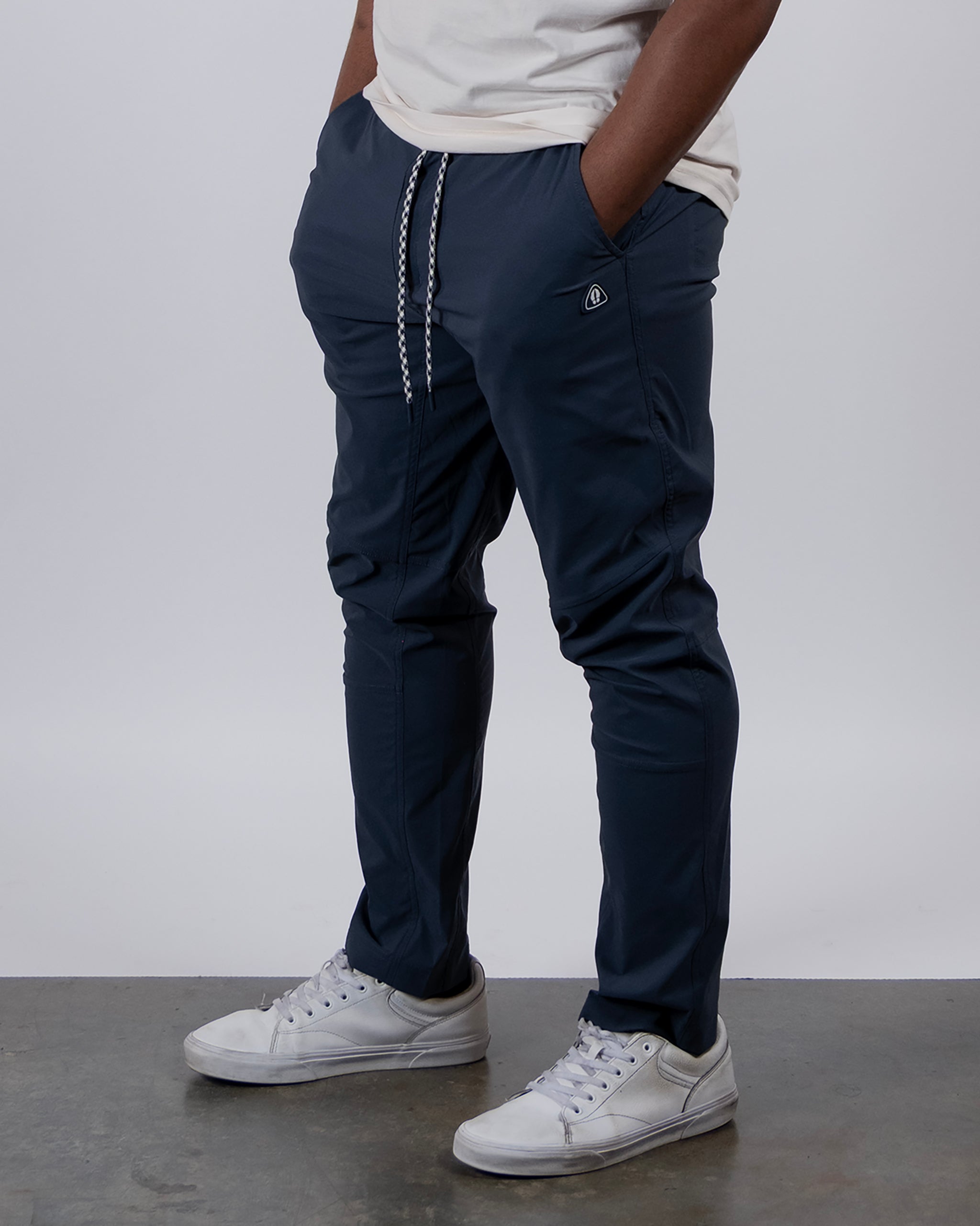 A person wearing a white t-shirt, BEARDEDGOAT NEW — ROVER™ Pant in navy blue with drawstrings, and white sneakers stands hands in pockets on a gray floor against a plain light background.
