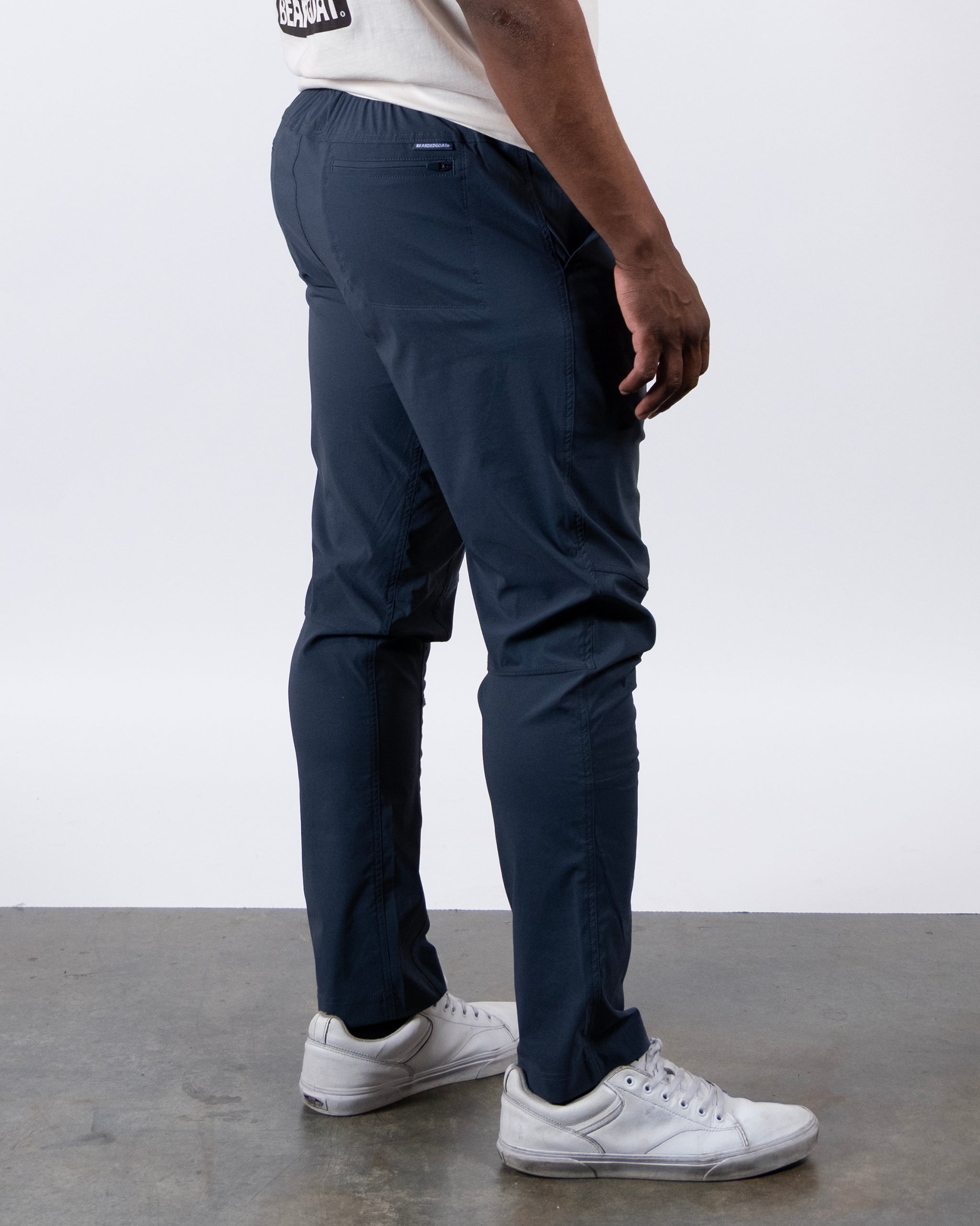 A person wearing BEARDEDGOAT's NEW — ROVER™ Pant in dark blue, a light T-shirt, and white sneakers stands sideways on a gray concrete floor with arms relaxed at their side.