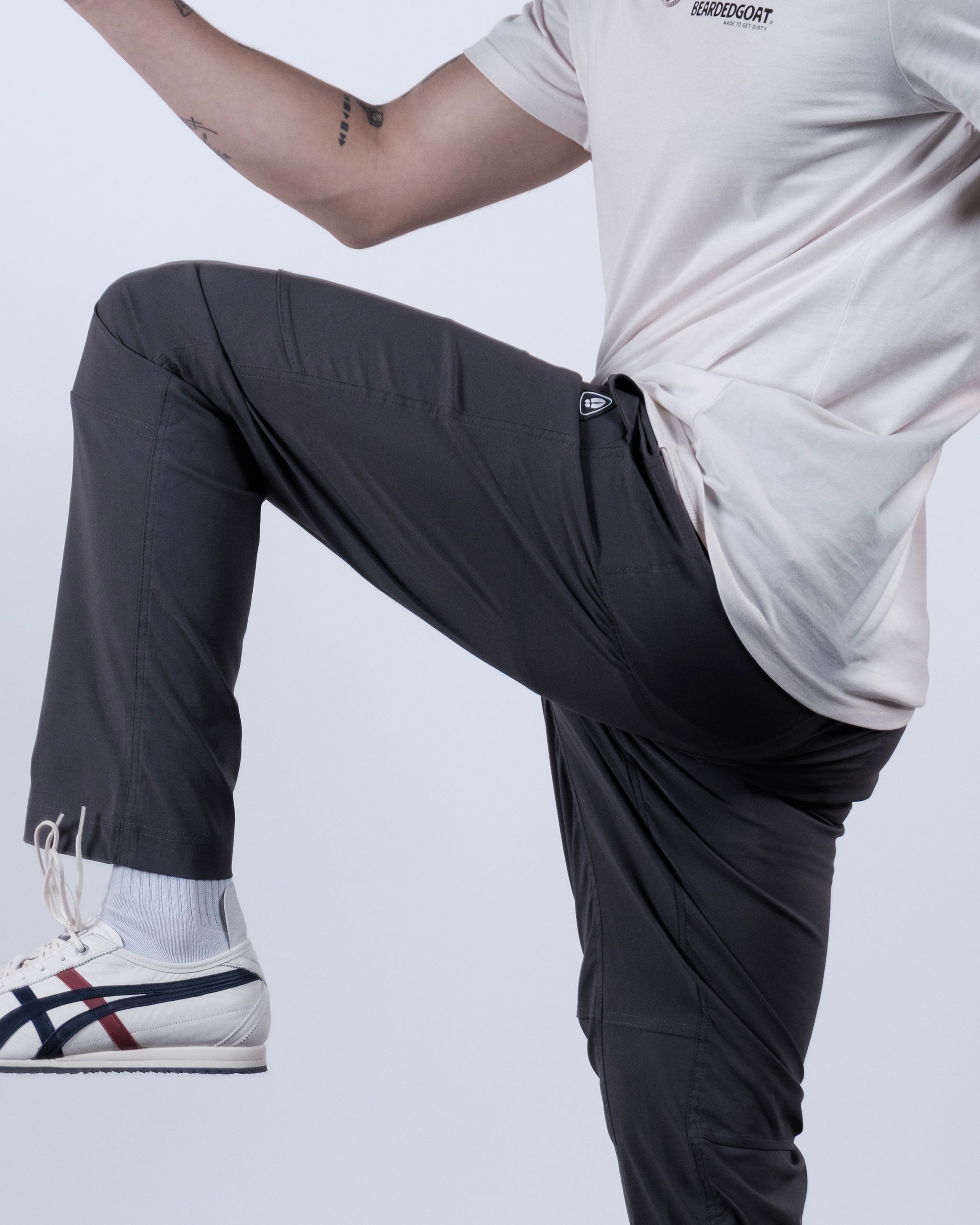 A person wears a beige t-shirt, BEARDEDGOAT NEW — ROVER™ Pant in dark gray, and white sneakers with blue and red stripes, raising one knee. Only their torso and legs appear against a plain background.