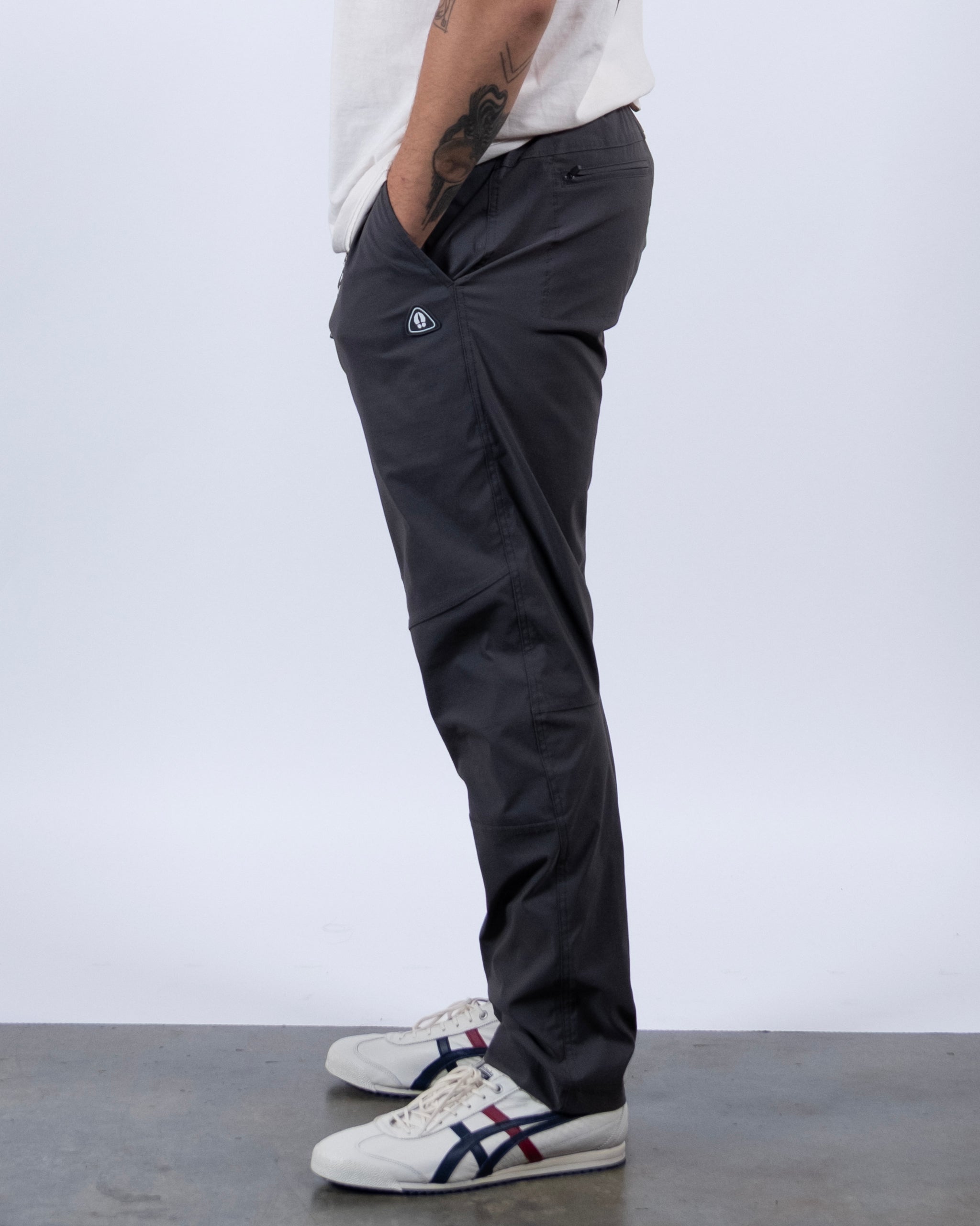 A person stands sideways in BEARDEDGOAT's NEW — ROVER™ Pant in dark gray, paired with a white t-shirt and white sneakers with navy and red stripes. One hand is in the pocket, showing part of a forearm tattoo. The background is plain and light-colored.