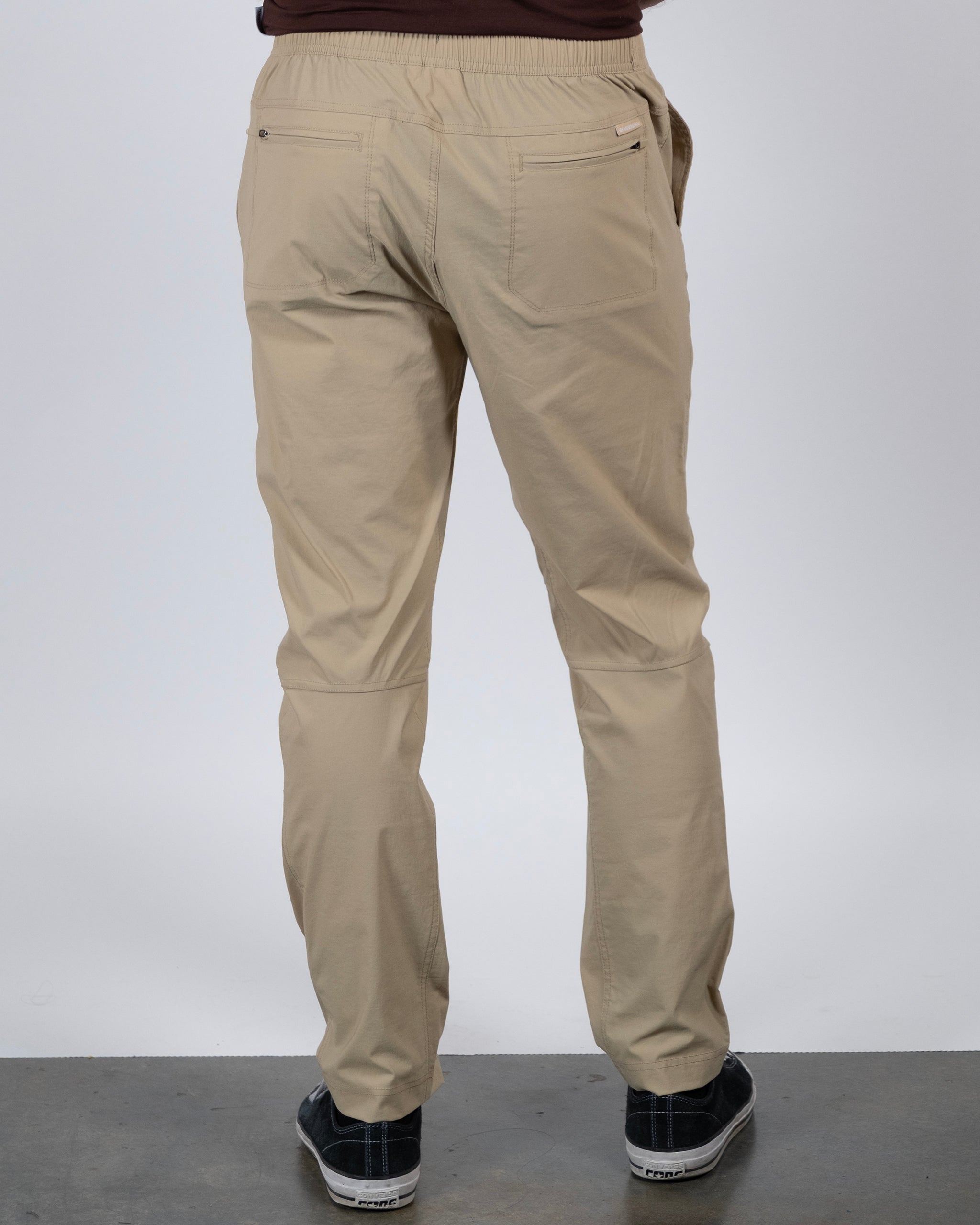 A person is standing on a gray floor facing away, wearing BEARDEDGOAT's NEW — ROVER™ Pant in beige, paired with black sneakers and a brown shirt. The image highlights the back view of the pants.