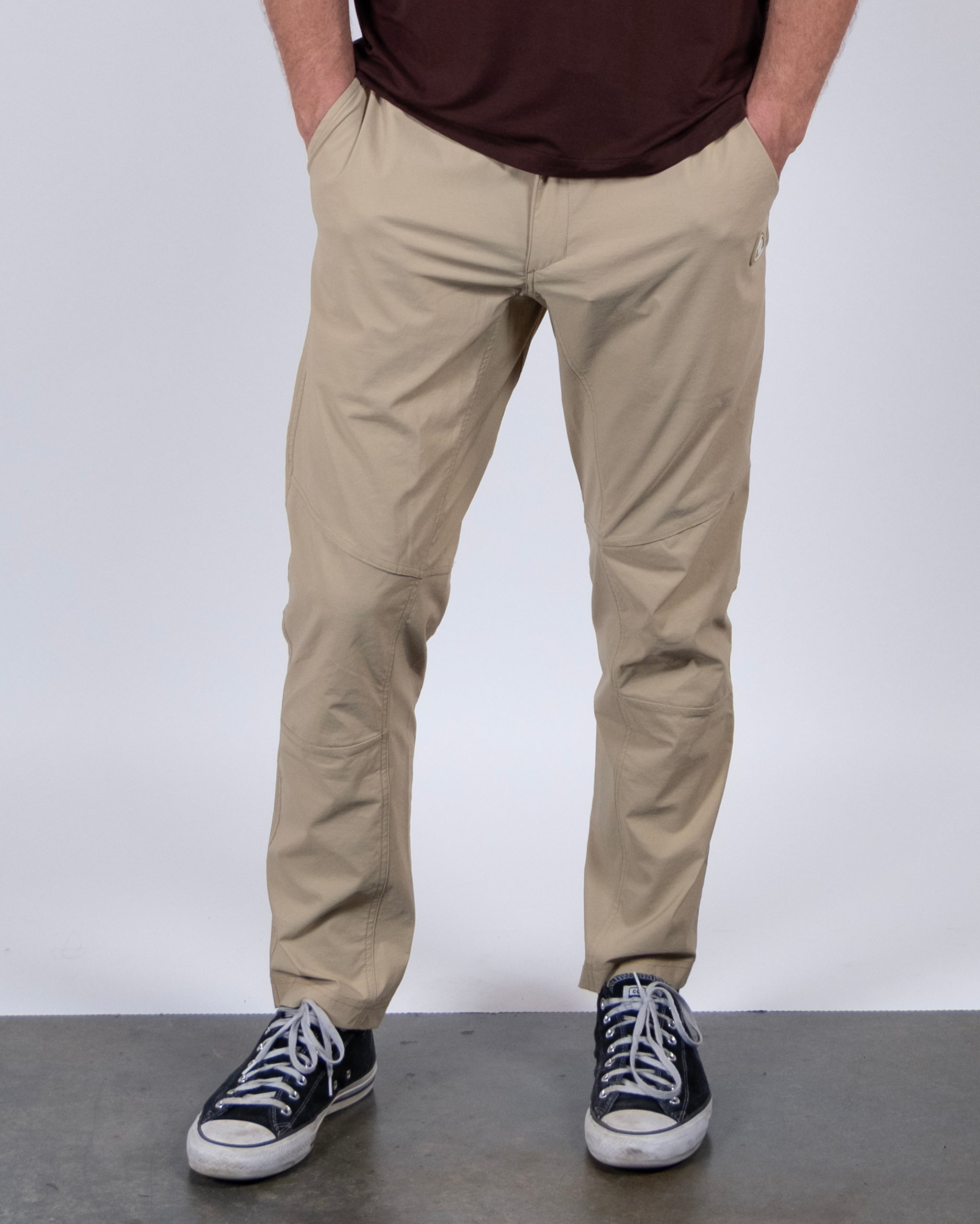 A person wears BEARDEDGOAT's NEW — ROVER™ Pant in light tan, a dark brown shirt, and black Converse sneakers, standing hands-in-pockets on a gray concrete floor with a plain white background.
