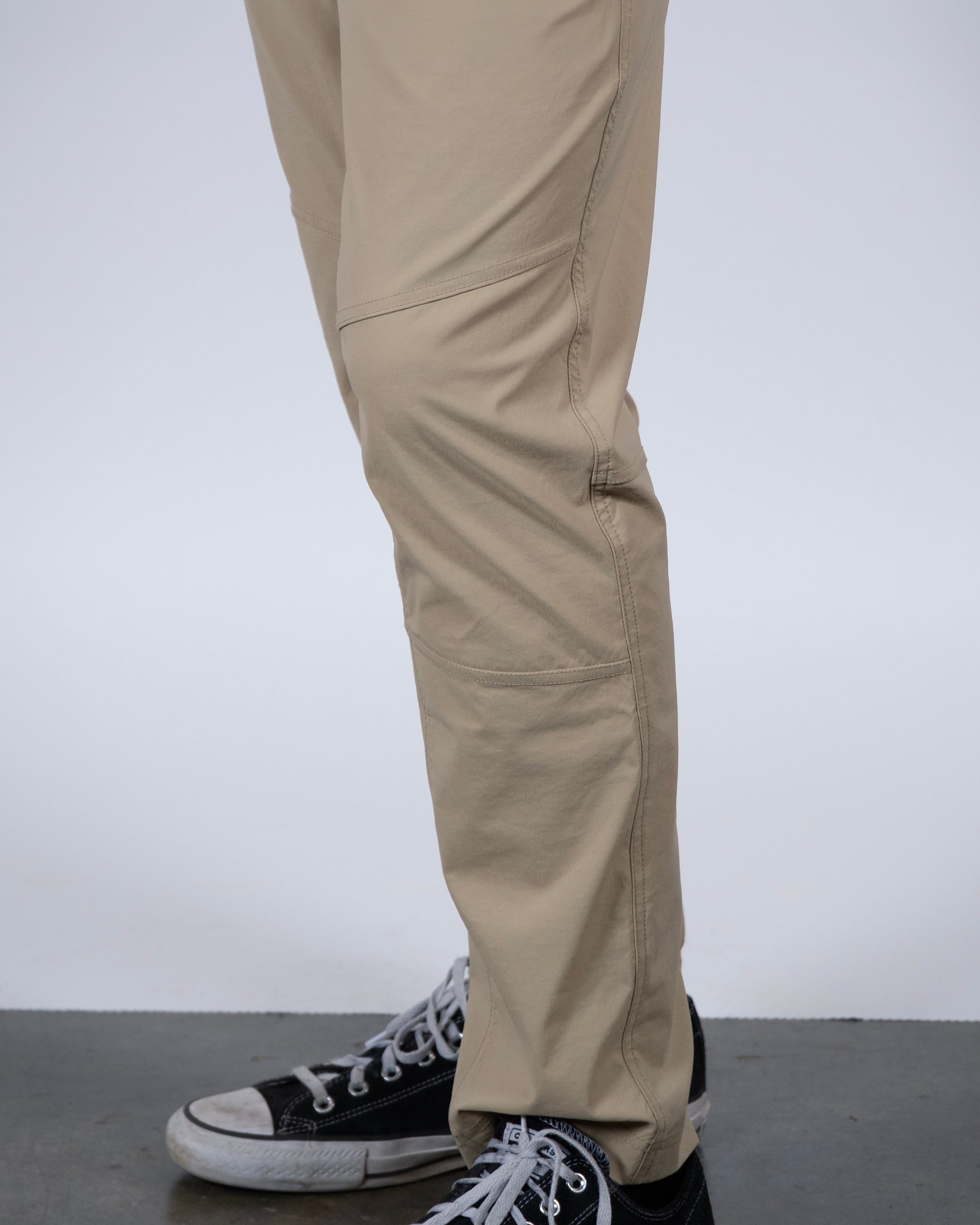 A person models the BEARDEDGOAT NEW — ROVER™ Pant in light tan with black-and-white sneakers, standing on a gray floor against a white background. The photo highlights their lower legs and footwear.