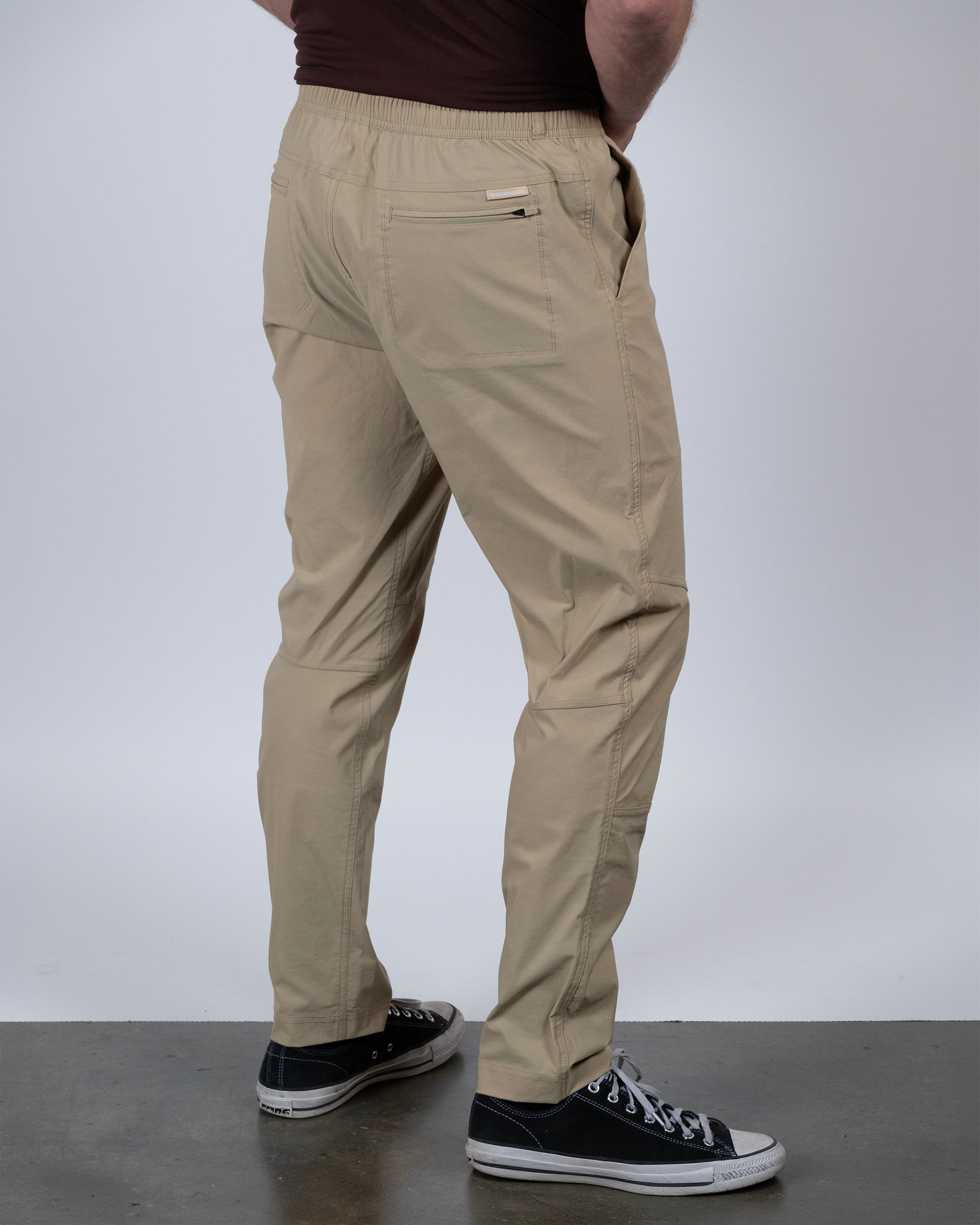 A person wearing BEARDEDGOAT's NEW — ROVER™ Pant and black sneakers stands on a gray floor, facing away with one hand in their pocket. The background is plain and light-colored.