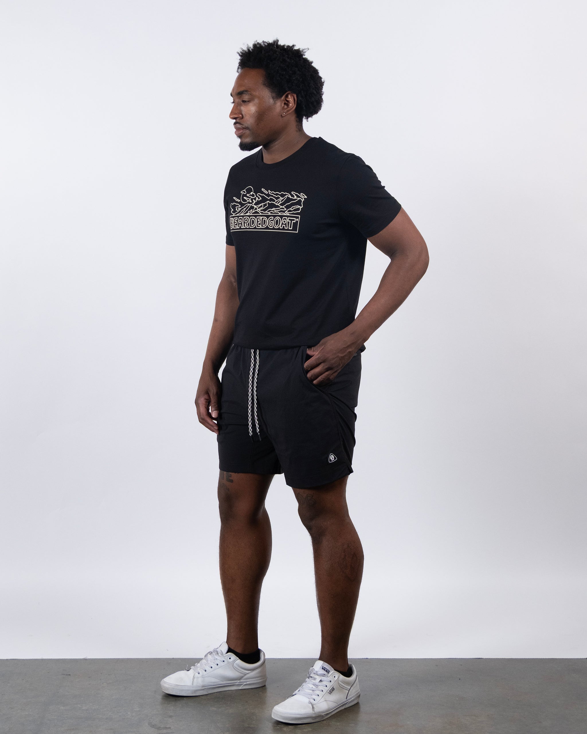A man stands against a plain white background wearing a black graphic T-shirt, BEARDEDGOAT Men's Rover Hybrid Short in black with a drawstring, and white sneakers. He looks to his left with one hand in his pocket.