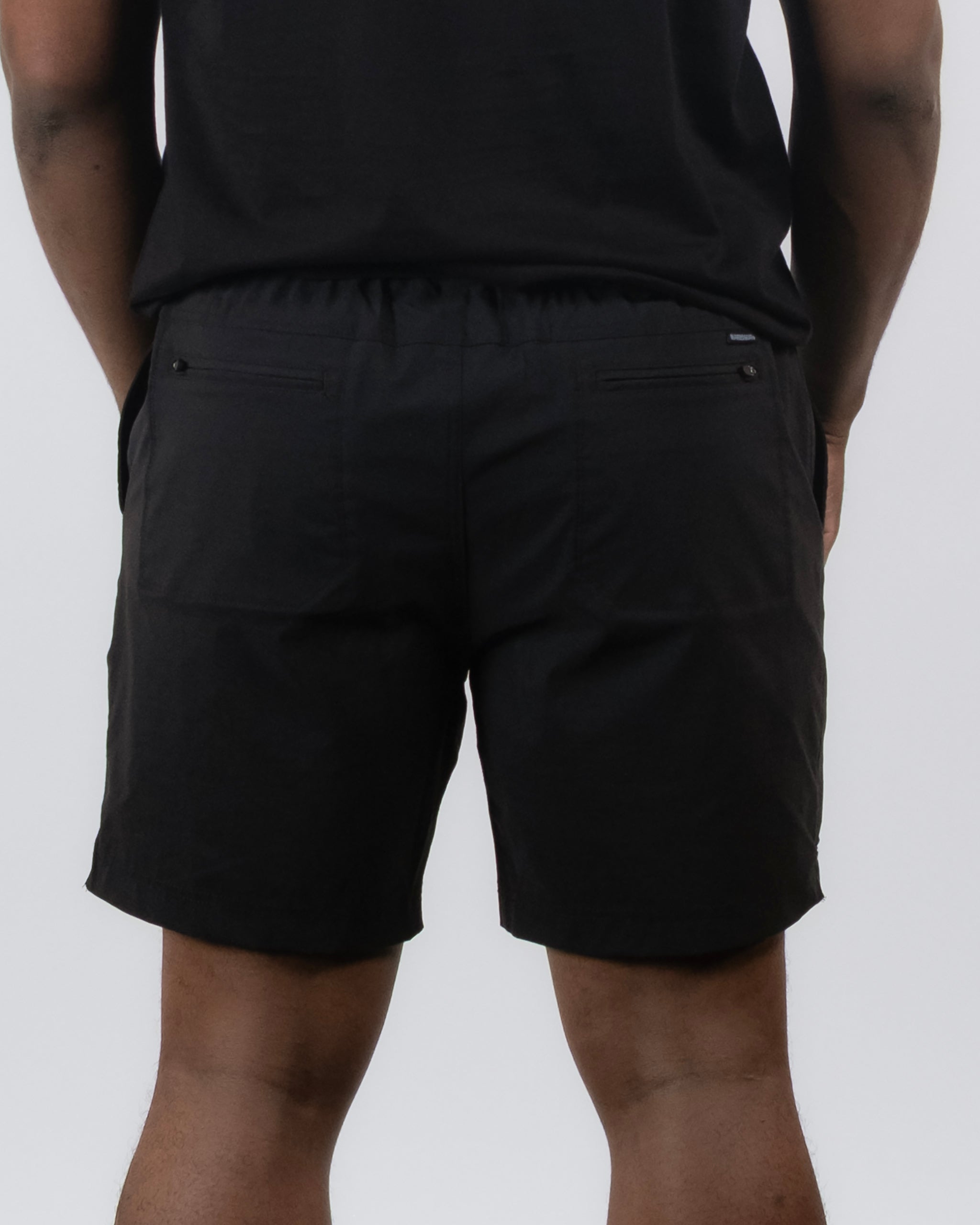 A person is shown from the back in BEARDEDGOAT Men's Rover Hybrid Short and a black T-shirt against a plain light background. The shorts feature stretch nylon fabric, back pockets, and an elastic waistband for comfort and versatility.