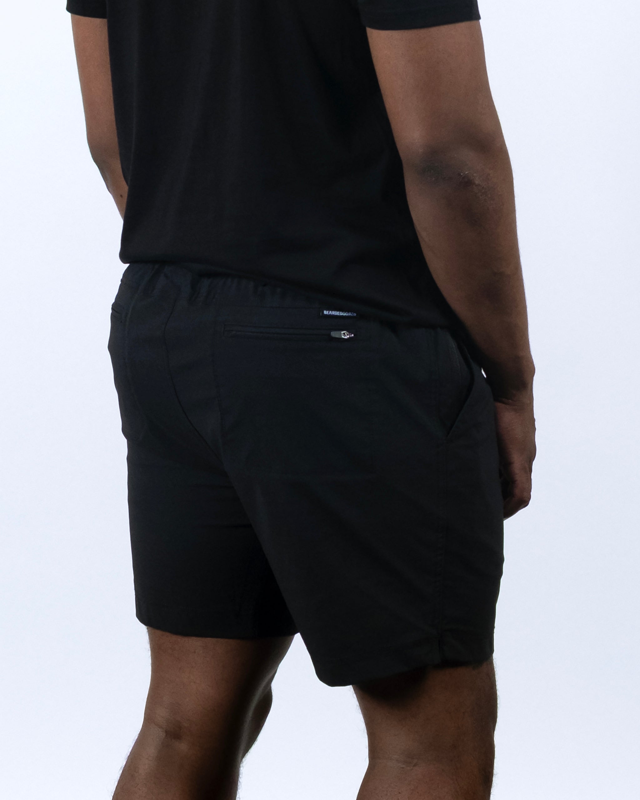 A person in a black BEARDEDGOAT Men's Rover Hybrid Short and black t-shirt stands facing away from the camera, left arm relaxed. The background is plain and light-colored.