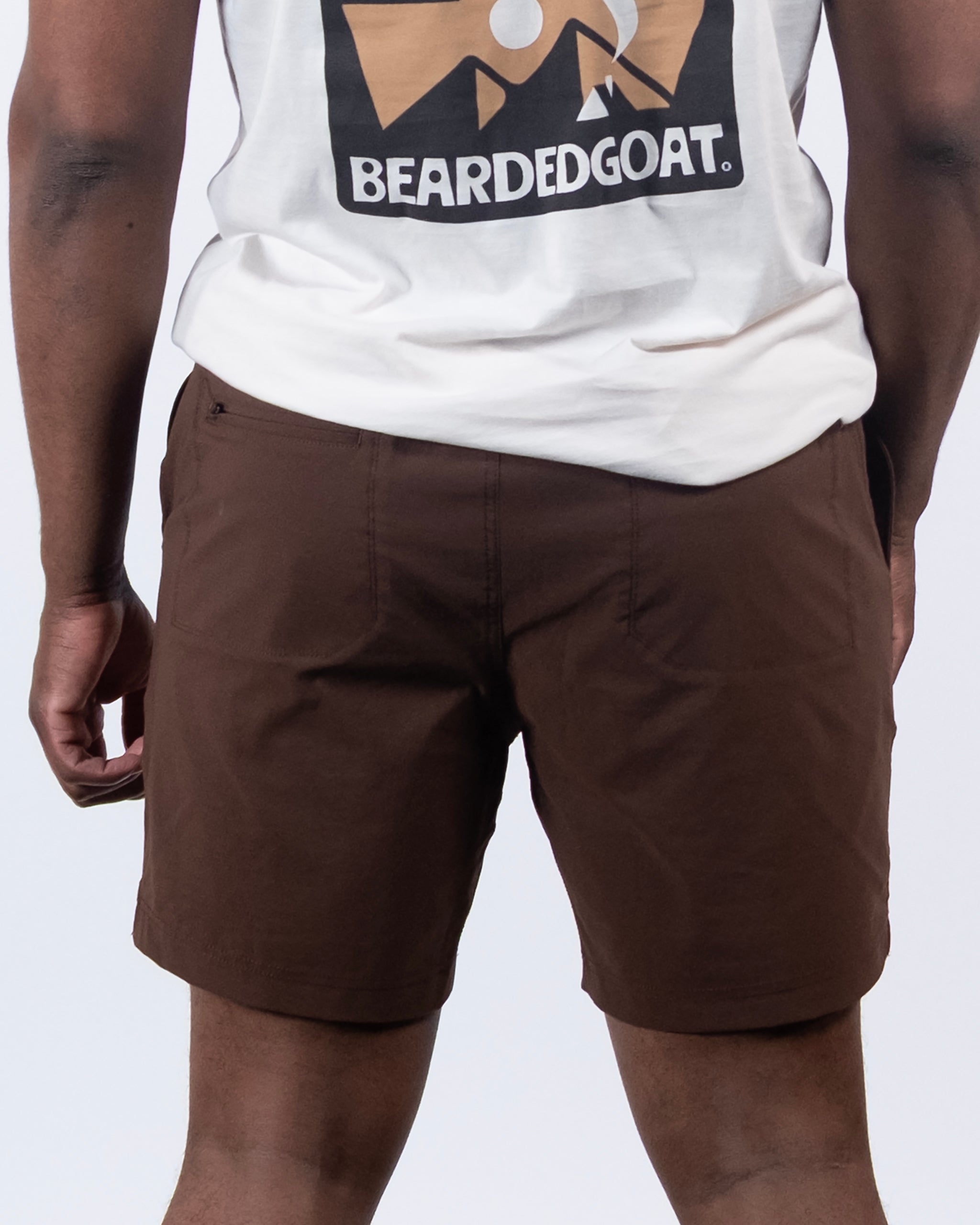A person shown from the back wears a white t-shirt with a mountain graphic and BEARDEDGOAT text, paired with BEARDEDGOAT Men's Rover Hybrid Shorts in brown.
