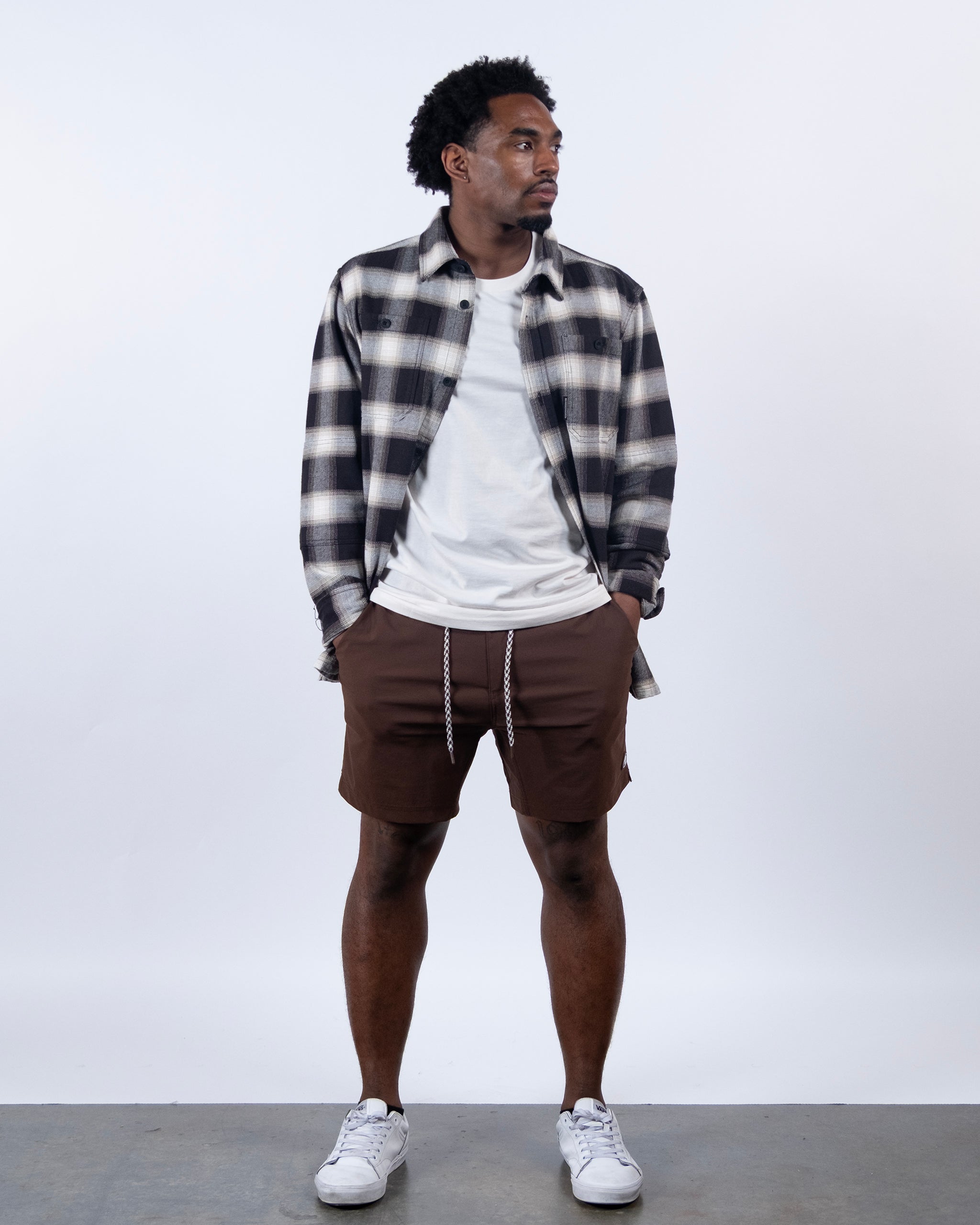 A man with short curly hair looks to his left, wearing a black-and-white plaid shirt over a white tee, BEARDEDGOAT Men's Rover Hybrid Short with drawstring, and white sneakers against a plain white background.