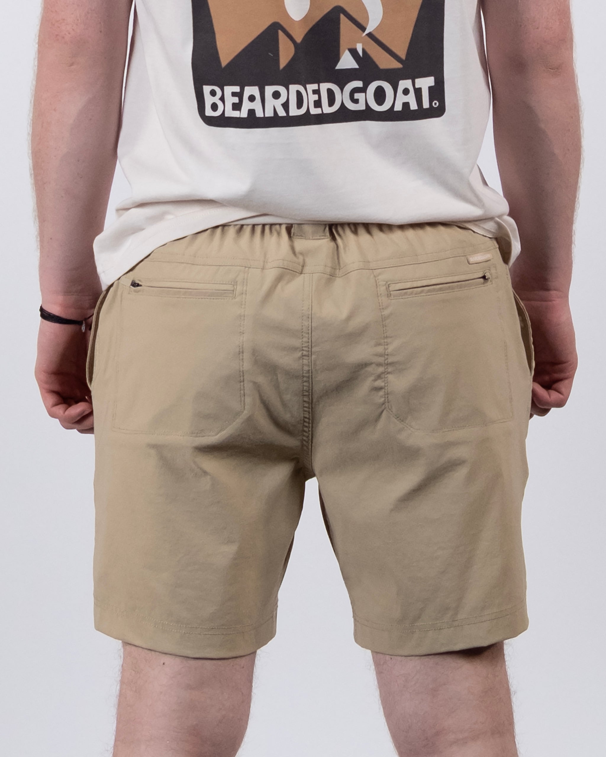 A person wears BEARDEDGOAT’s beige Men’s Rover Hybrid Short, featuring stretch nylon, elastic waistband, and back pockets, paired with a white BEARDEDGOAT graphic tee. The rear view emphasizes the shorts’ comfort and versatility.