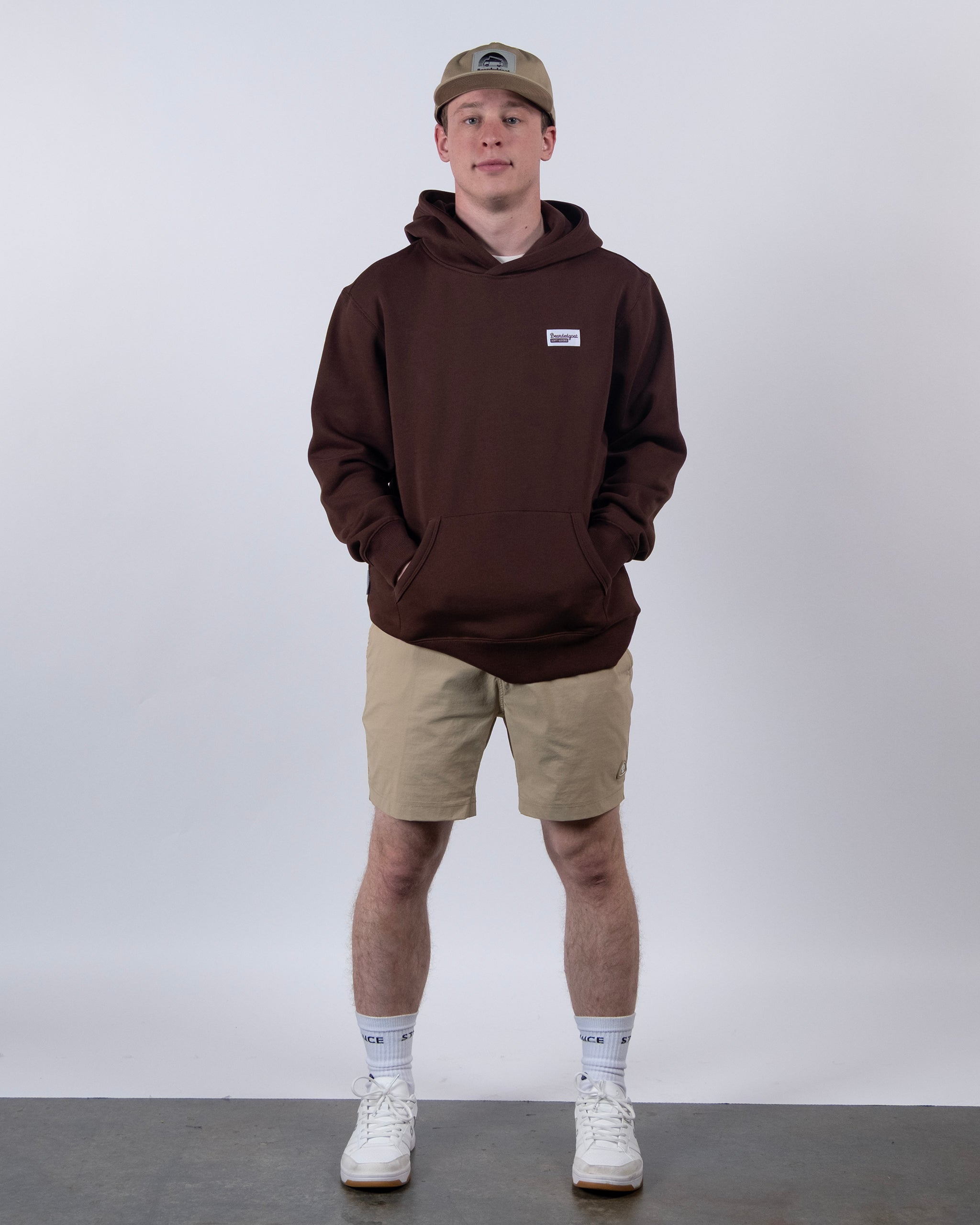 A person stands facing forward against a light background, wearing a brown hoodie, beige BEARDEDGOAT Men's Rover Hybrid Short, a beige cap, white crew socks, and white sneakers. Hands are in the hoodie pocket.