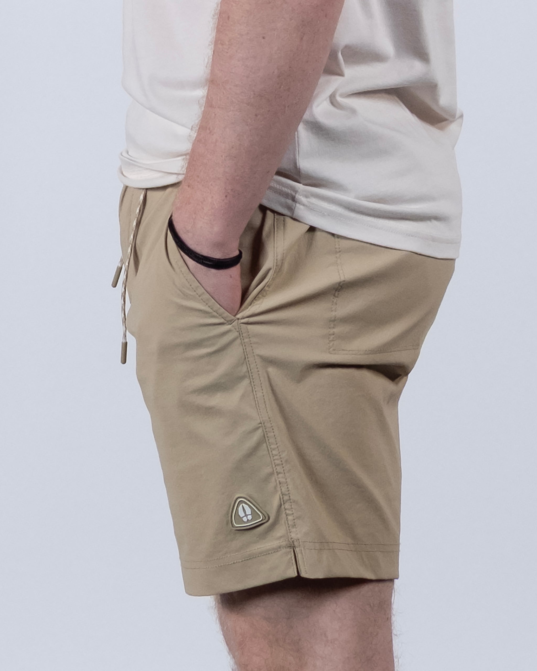 A person in a beige T-shirt and BEARDEDGOAT Men's Rover Hybrid Short with hands in pockets, seen from the side against a plain light background. The versatile shorts display a small triangular logo by the hem.